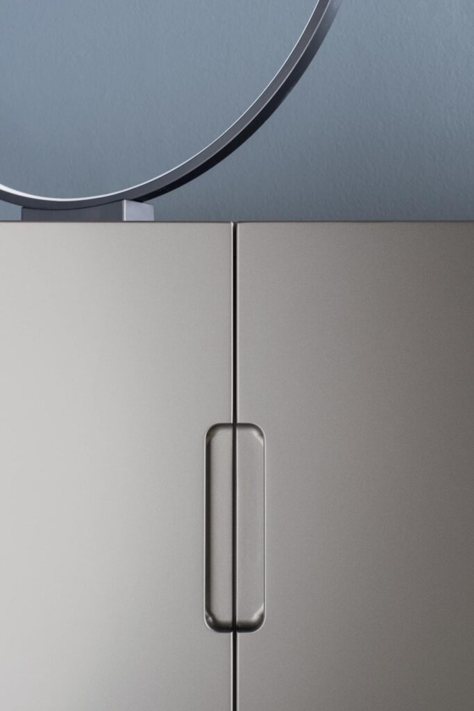A close-up vertical shot of a modern, light-gray cabinet. The image focuses on the seam between the two doors, highlighting the sleek, recessed vertical handle. A thin, circular piece of metal from an object resting on top of the cabinet is visible at the very top of the frame against a blurred blue background.