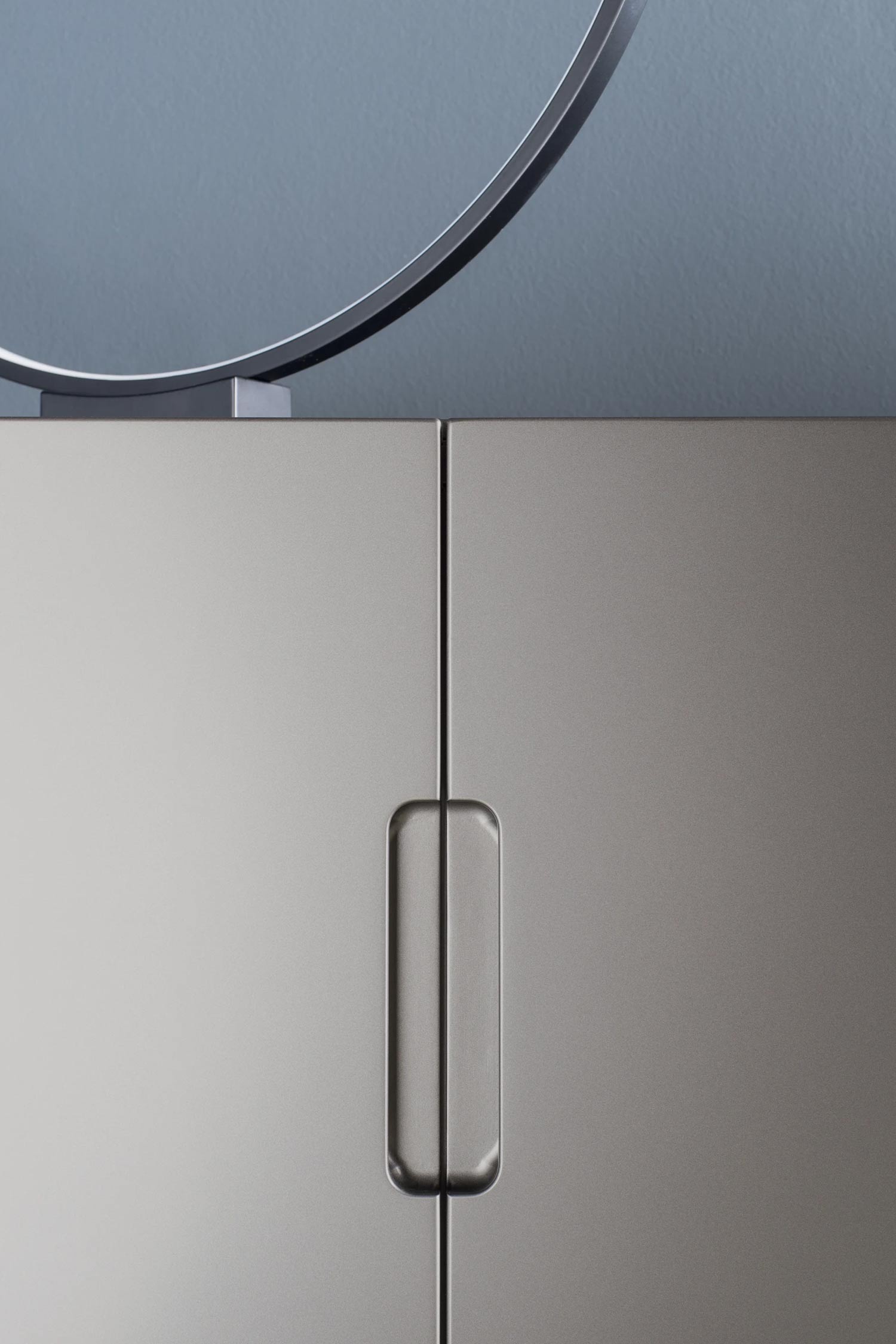 A close-up vertical shot of a modern, light-gray cabinet. The image focuses on the seam between the two doors, highlighting the sleek, recessed vertical handle. A thin, circular piece of metal from an object resting on top of the cabinet is visible at the very top of the frame against a blurred blue background.