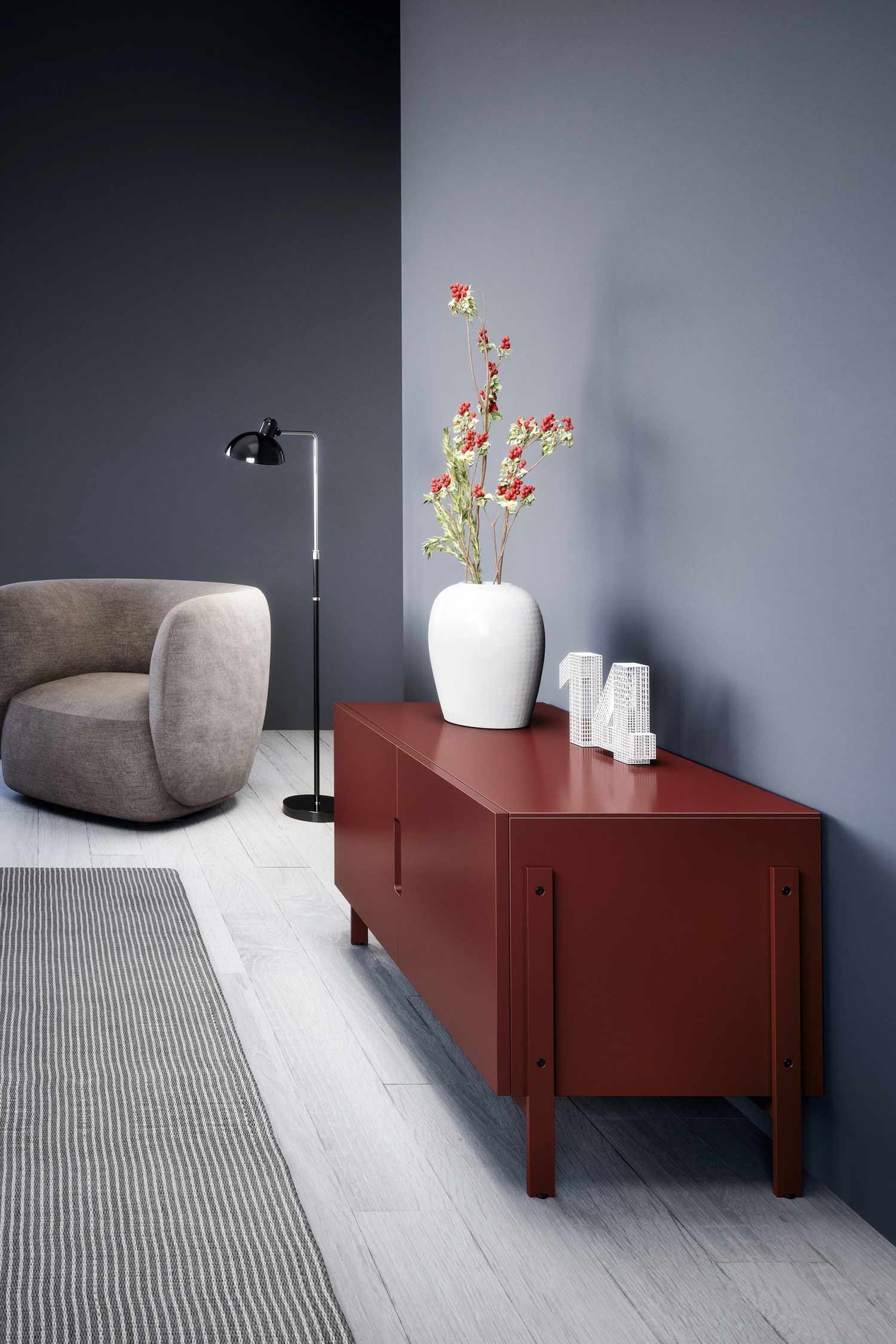 A cozy, modern living space with a dark gray accent wall and a lighter gray wall meeting in the corner. In the foreground, a long, deep red console table stands against the corner, adorned with a white vase holding red berries and green leaves, and a white decorative object shaped like the number "14." To the left of the console, a plush, light brown armchair with a rounded back is partially visible, next to a black floor lamp with a curved arm and a round shade. The floor is light wood with a striped rug extending from the bottom left.