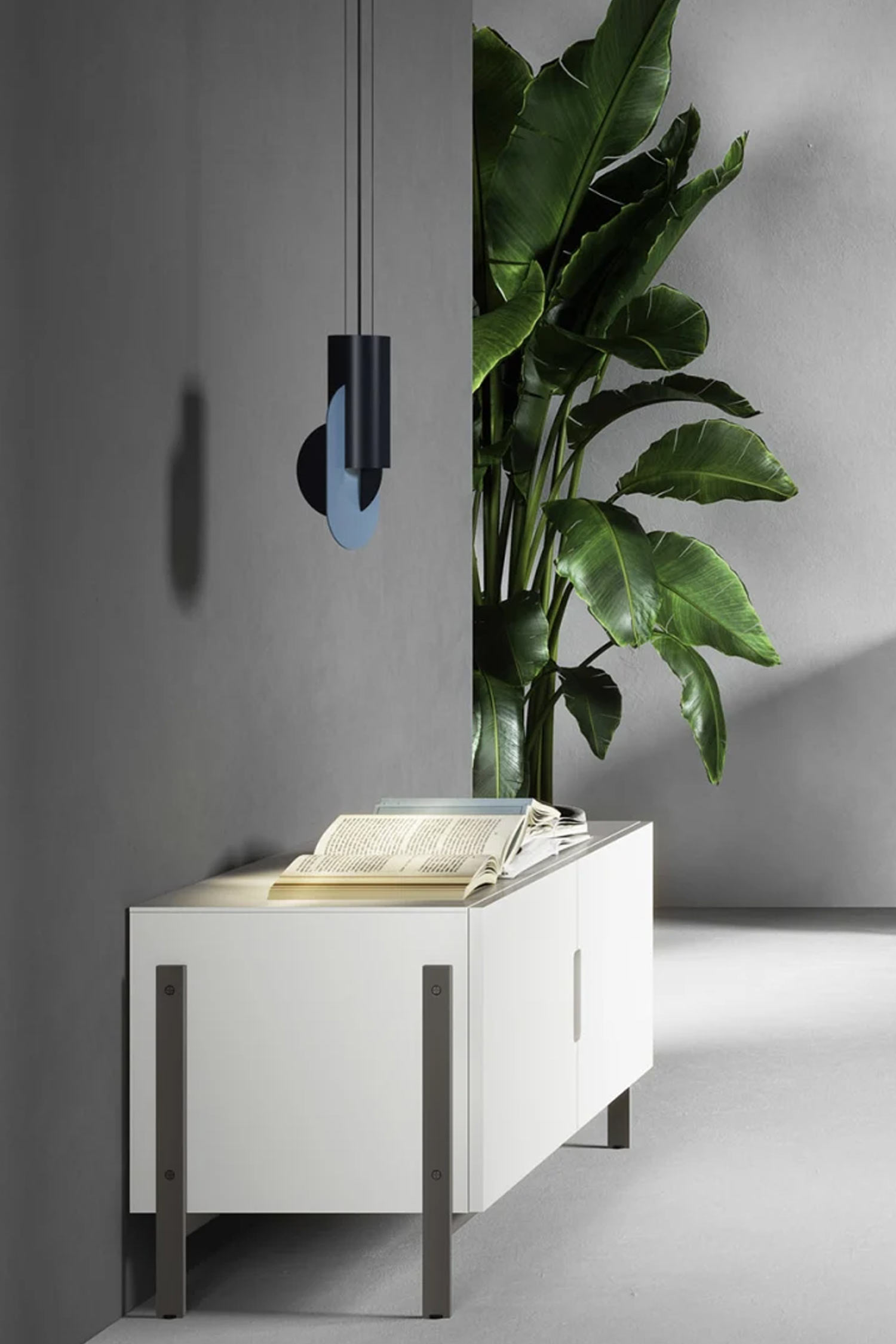 A low-angle shot of a white two-door cabinet with a modern design, resting on four gray metal legs. On top of the cabinet, an open book and other reading materials are visible. A large, green, tropical plant stands behind the cabinet. A unique, sculptural pendant light with dark blue and lighter blue oval shapes hangs from the ceiling to the left of the cabinet, casting a shadow on the dark gray wall behind it. The floor is light gray.