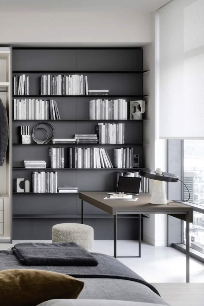 A close-up of a modern home office space within a bedroom. The focus is on a sleek, dark brown desk with black metal legs. On the desk are a laptop and a unique white stone lamp with a black rectangular shade. The desk is placed against a large, dark gray wall unit with multiple shelves filled with books and various decorative objects. In the foreground, a bed with gray bedding is partially visible, and a small, textured, upholstered stool sits near the desk.