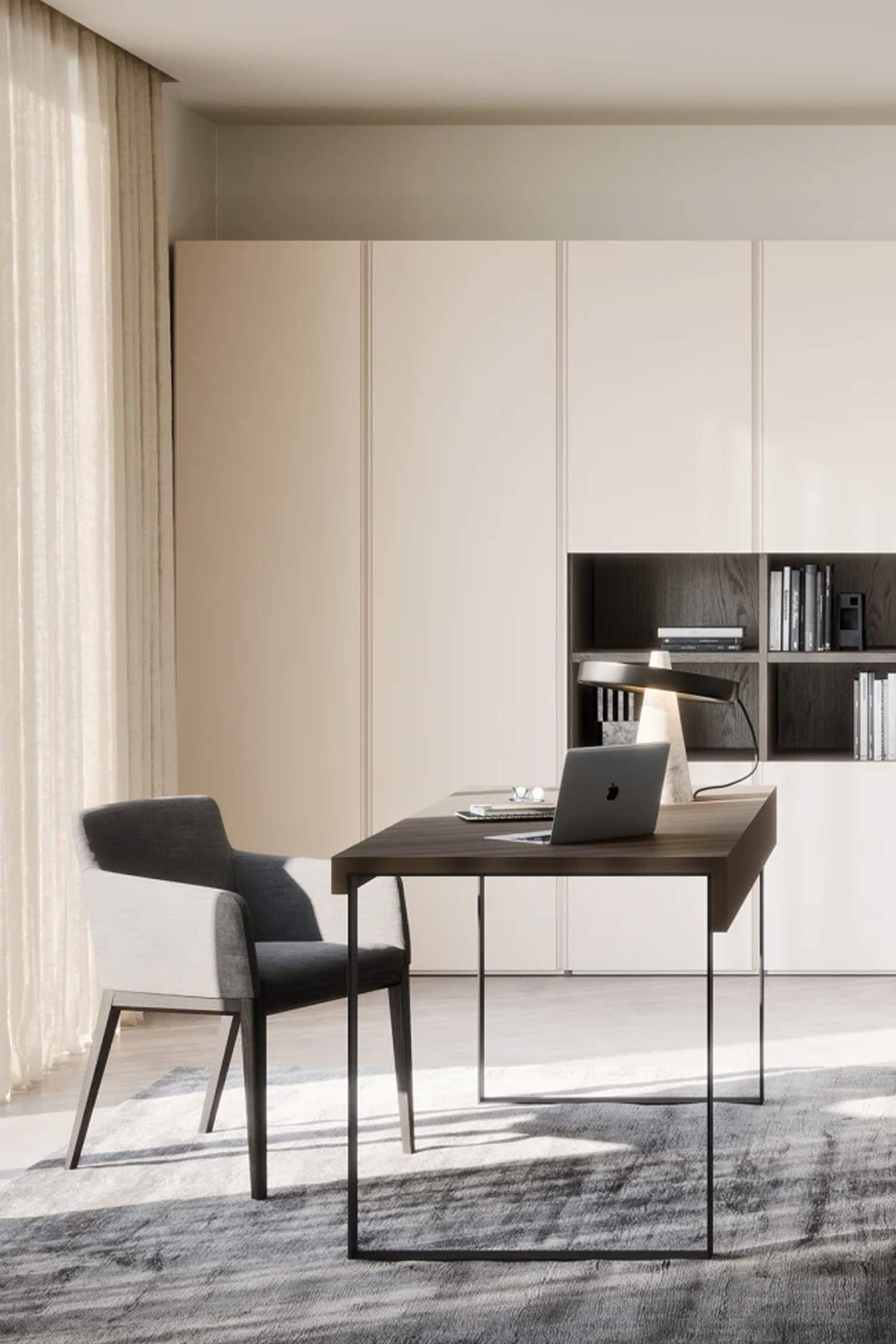 A home office with a minimalist aesthetic. In the center, a dark wood desk with thin black metal legs holds a laptop and a unique black lamp with a white marble base. To the left is a modern chair with a dark gray seat and a light gray back. In the background, large built-in cabinets in a light cream color feature a central section with dark gray open shelving filled with books. The room is bathed in natural light from a large window on the left, which is covered by a sheer curtain. The floor is covered with a large gray rug.