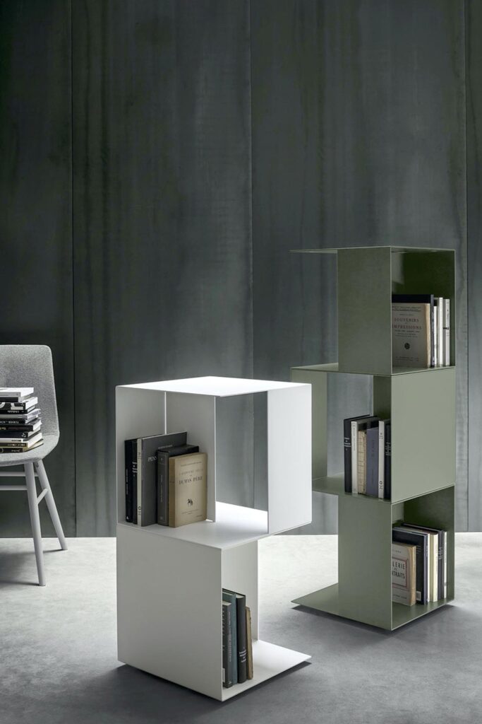 An interior room with a gray concrete floor and a dark gray, textured wall. In the foreground, a modern, modular bookshelf made of metal cubes is shown. On the left, two white cubes are stacked on their side with books inside. On the right, a stack of three olive green cubes holds books. A gray upholstered chair is visible on the far left.