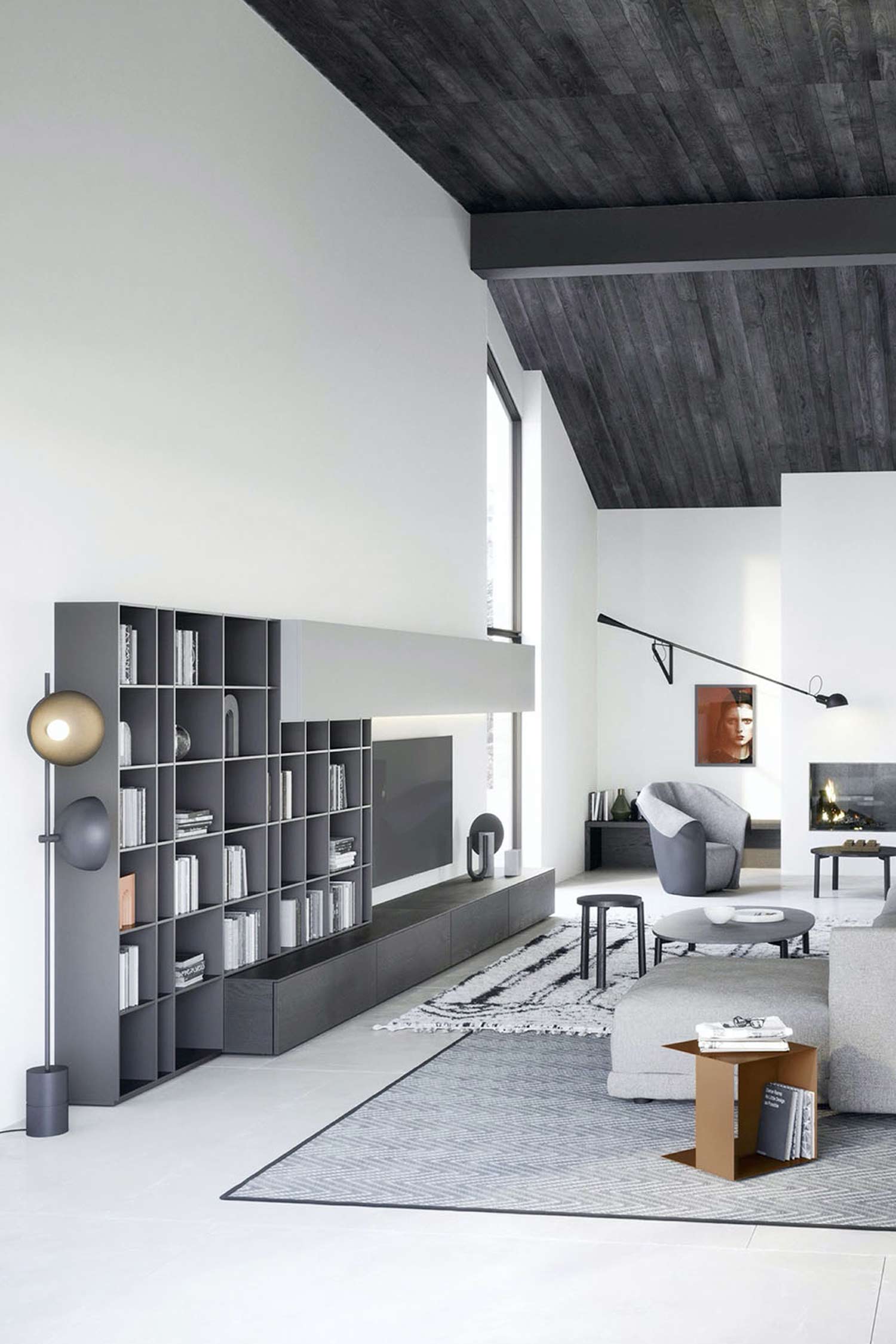 A spacious, modern living room with a vaulted ceiling clad in dark wood. A large, dark gray modular shelving unit with a built-in media console and space for a television is against the wall on the left. The shelves are filled with books. In the center of the room, there's a gray sectional sofa, a small gray coffee table, and a matching side table. A large gray rug with a geometric pattern covers the light-colored floor. A glimpse of a fireplace and a gray armchair is visible in the background.