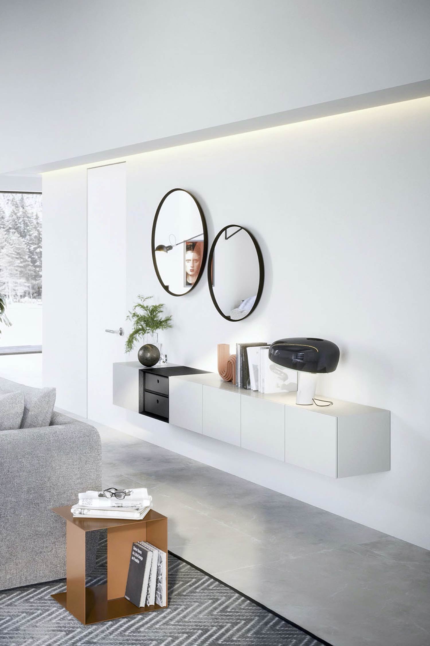 A bright, minimalist living room with a white floating media console mounted on a white wall. The console has several white cabinets and a section with two black drawers. Decorative items on top include a leafy plant, abstract vases, and a black, modern desk lamp. Two oval-shaped mirrors with dark frames are mounted on the wall above the console. The floor is tiled in light gray, and a bronze-colored side table with books is in the foreground.