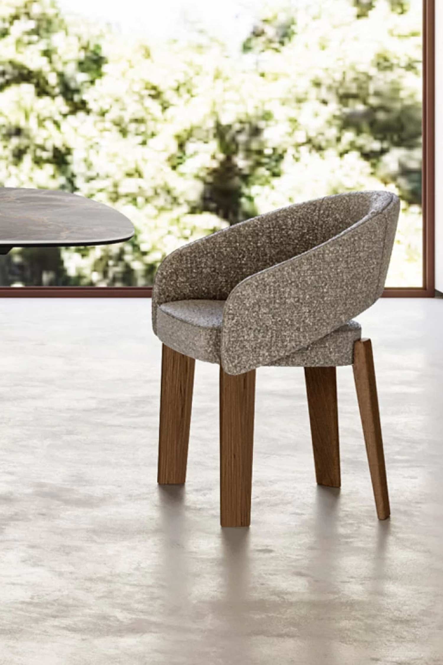 A close-up view of a dining chair with textured gray fabric upholstery and four dark wooden legs. The chair is placed on a light-colored floor, with a dining table and a large window with trees outside in the blurred background.