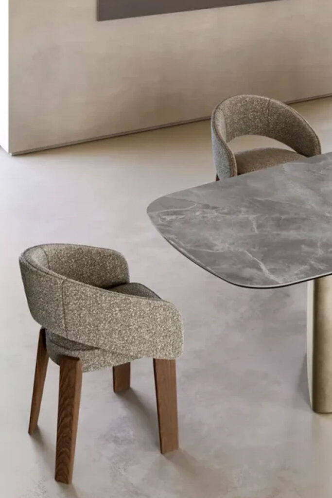 An angled, close-up view of two dining chairs with textured gray fabric upholstery and dark wooden legs. One chair is in the foreground, and the other is partially visible behind it, next to a dining table with a gray marble-like top.