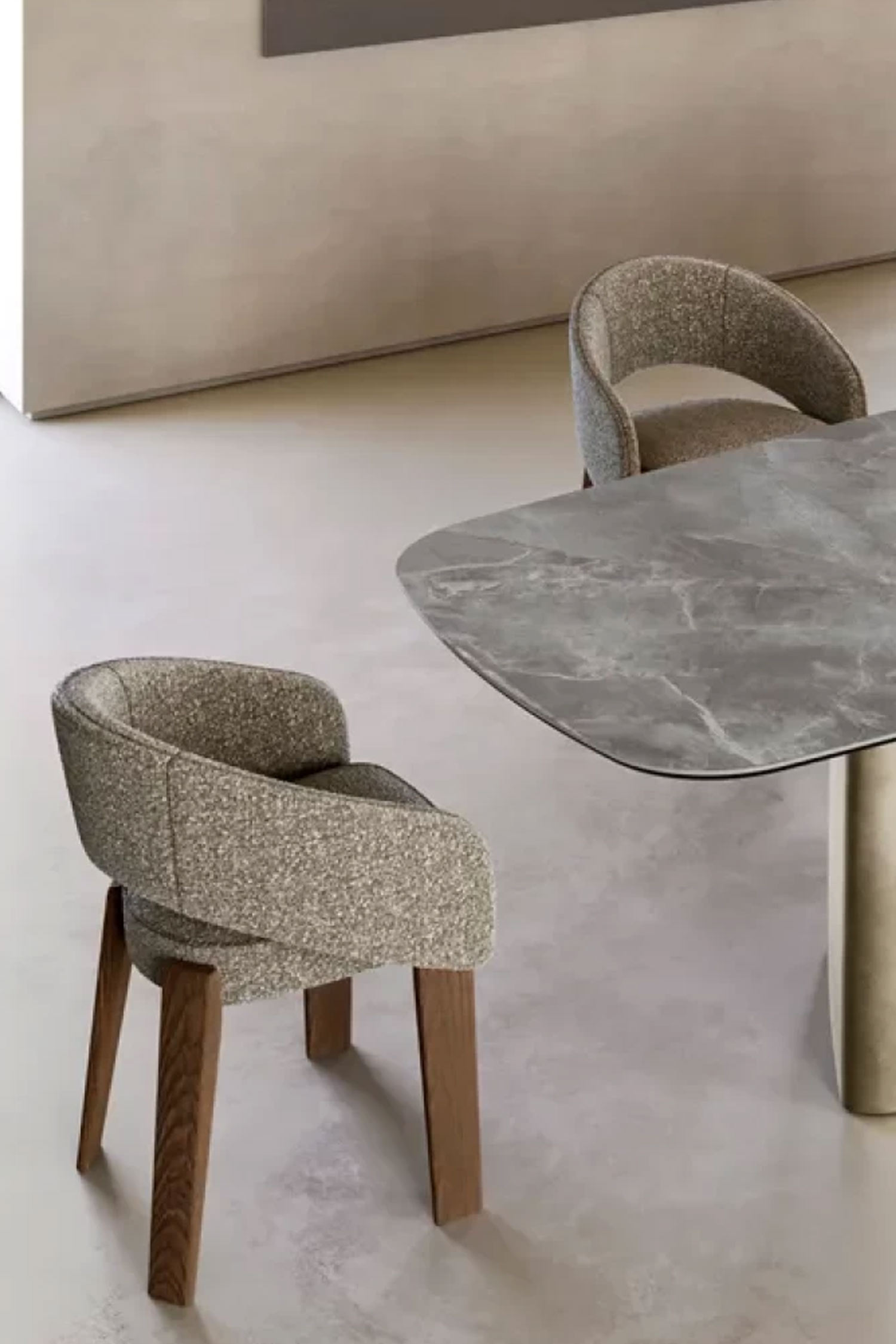 An angled, close-up view of two dining chairs with textured gray fabric upholstery and dark wooden legs. One chair is in the foreground, and the other is partially visible behind it, next to a dining table with a gray marble-like top.