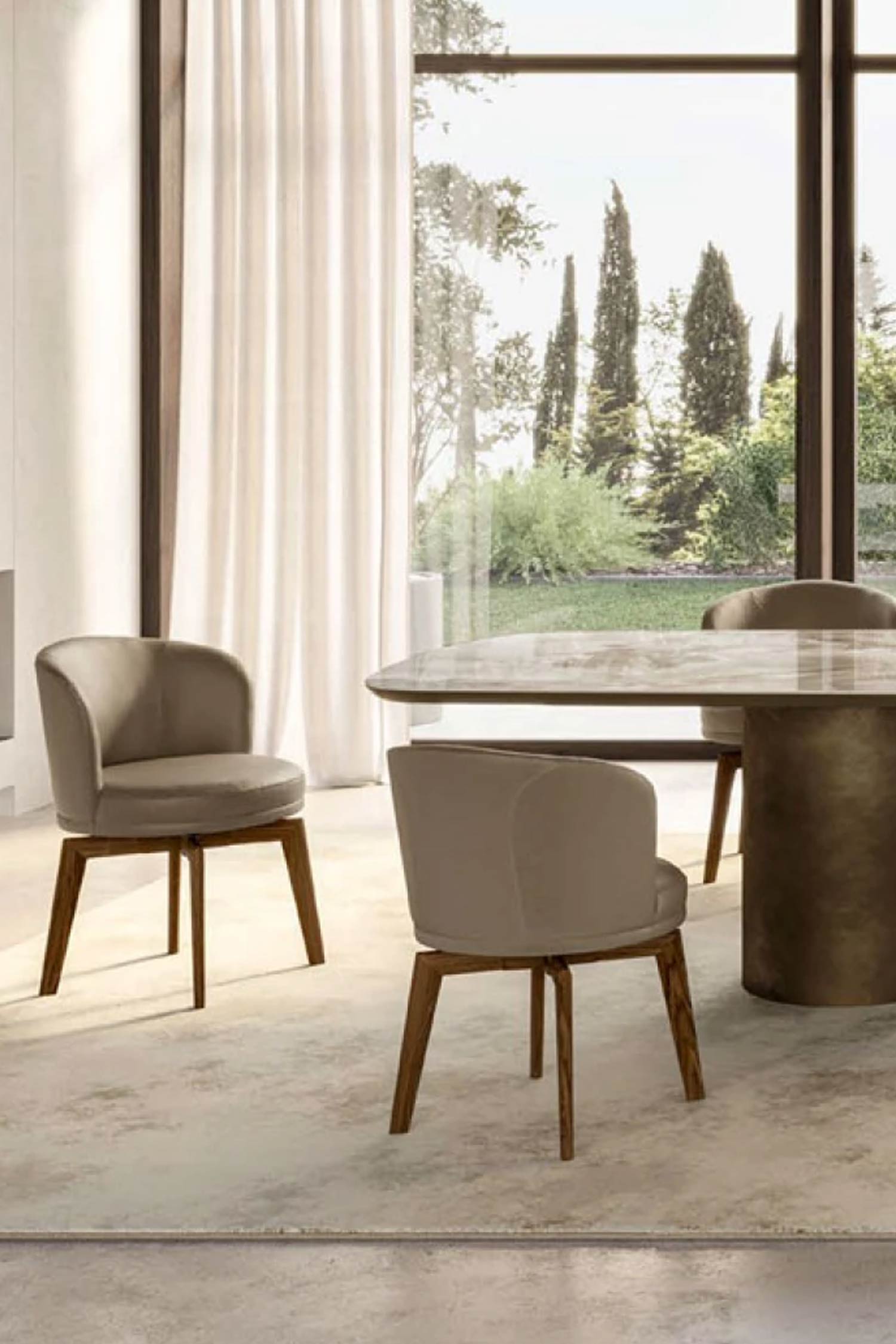 A detailed, slightly elevated view of two modern dining chairs with light brown upholstery and wooden legs. They are positioned on a light-colored rug next to a large dining table with a thick, cylindrical bronze base. The room has large windows with sheer white curtains, offering a view of a lush green lawn and cypress trees outside.