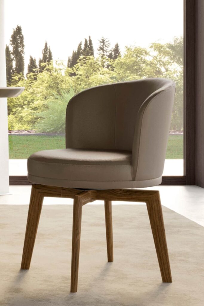 A close-up of a modern dining chair with light brown upholstery and a unique wooden base. The chair's seat and back are a single, continuous curve, and the legs splay outwards from a central point, creating a star-like shape. The image is taken from a low angle, showing the chair on a rug with a bright window and greenery in the background.