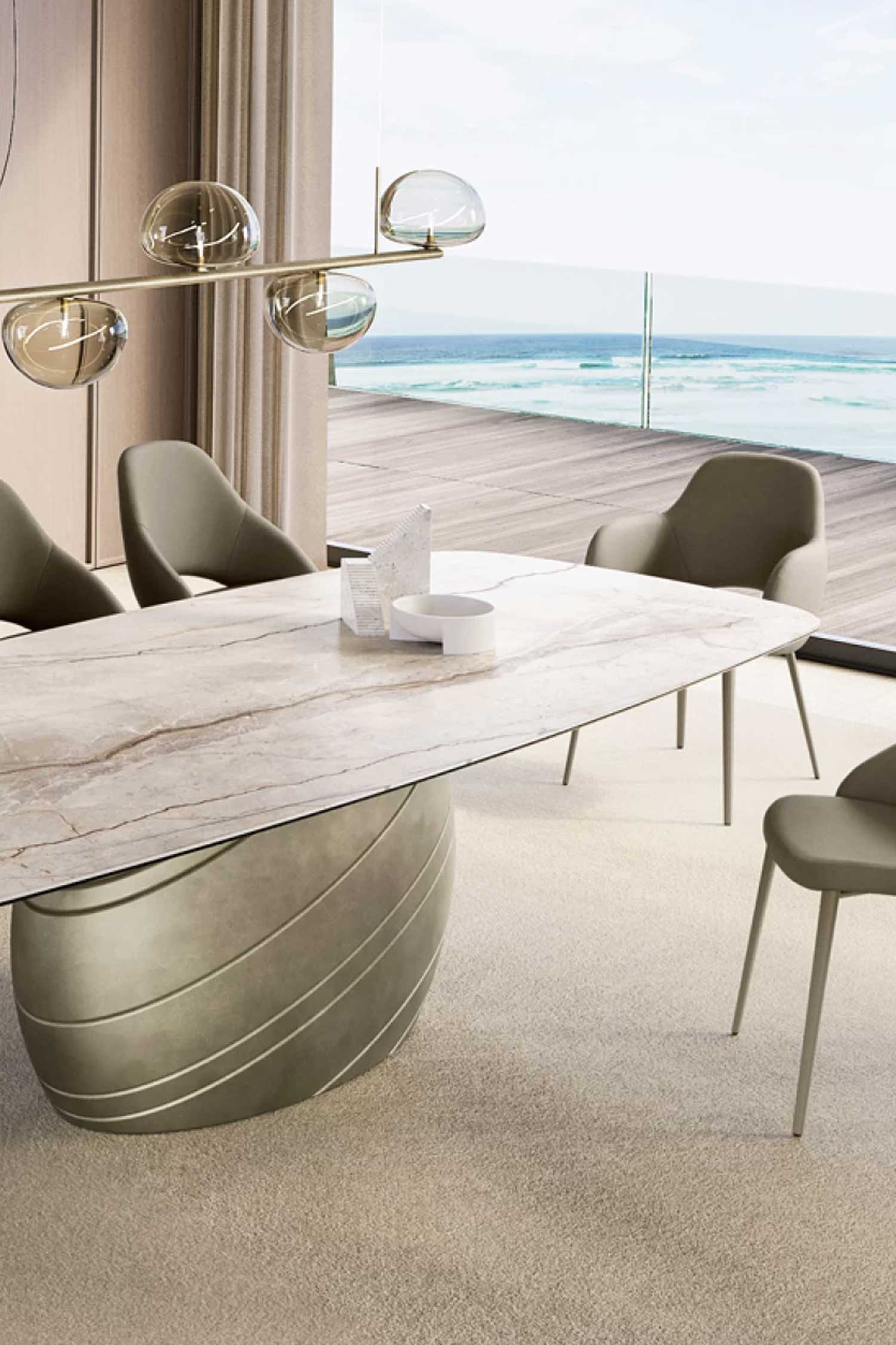 Close-up of the Giove luxury dining table featuring its marble top and a large, rounded, striated metallic base in a dining room with an ocean view. The setting includes minimalist taupe dining chairs and a linear pendant light.