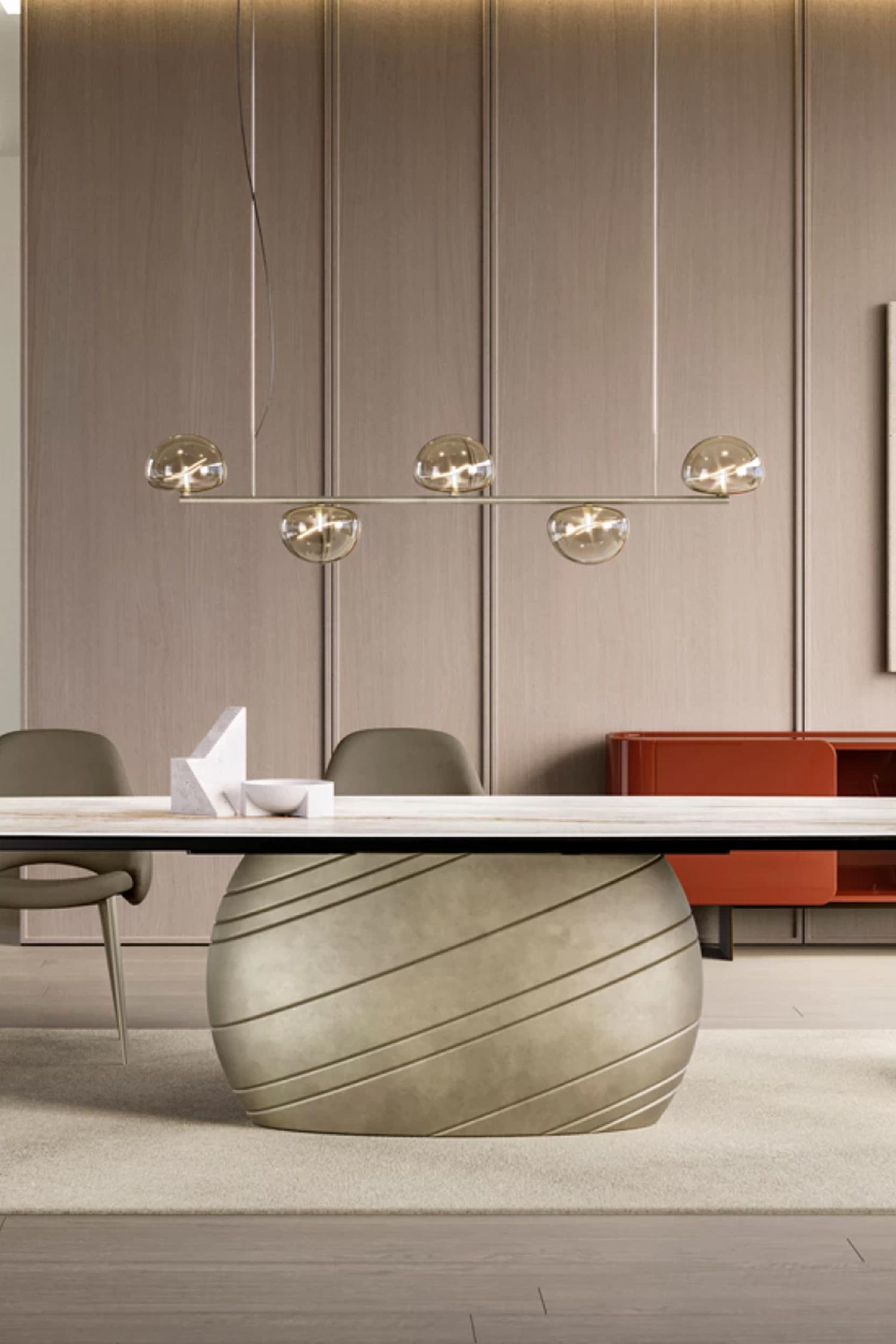 Detail shot of the Giove luxury dining table's massive, spherical base with diagonal metallic grooves. The table is shown in a modern dining room with wood-paneled walls and a linear pendant light featuring four glowing glass globes overhead.