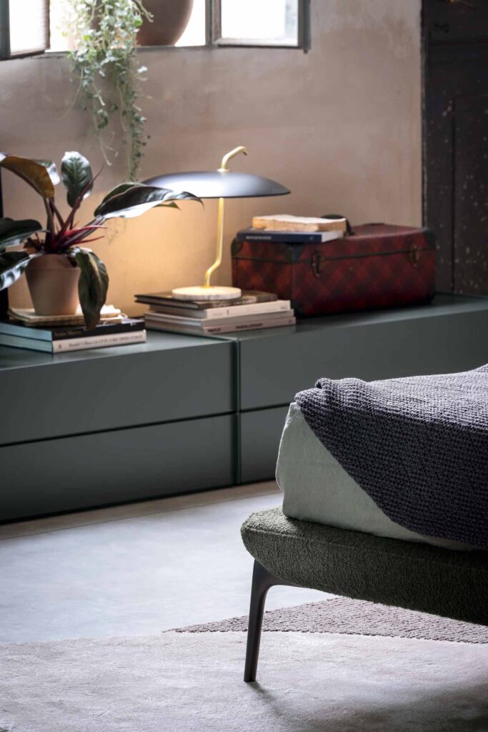 A close-up detail of a bedroom showing the foot of an upholstered green bed with a gray blanket, and a low, matte green drawer unit. The unit serves as a bedside table, holding books, a planter, a lamp, and a vintage-style red trunk.