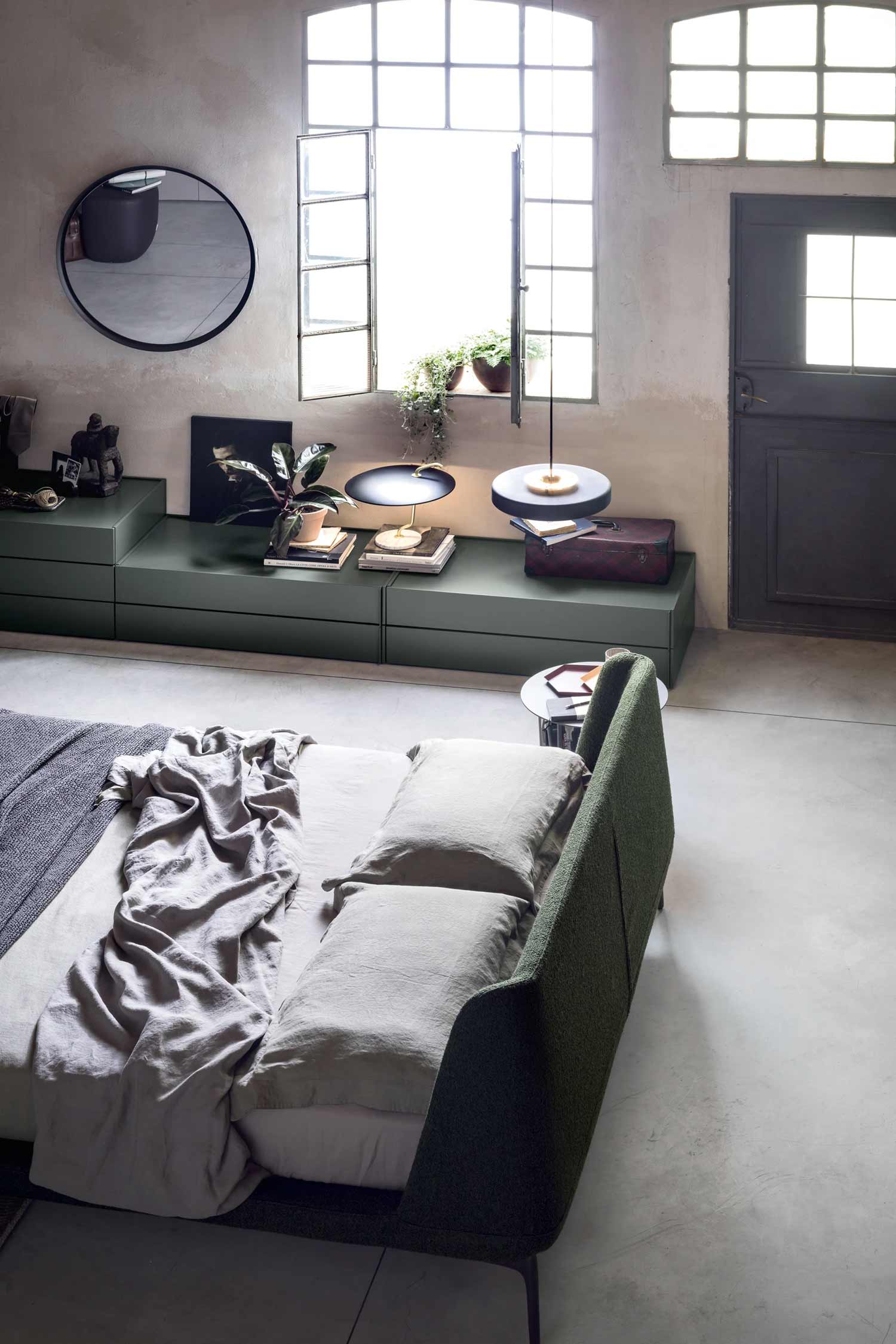 An industrial-style bedroom with a green upholstered bed, a round mirror, and a long, low matte green drawer unit running along a textured wall beneath a multi-pane window. The unit holds books, plants, and modern lamps.