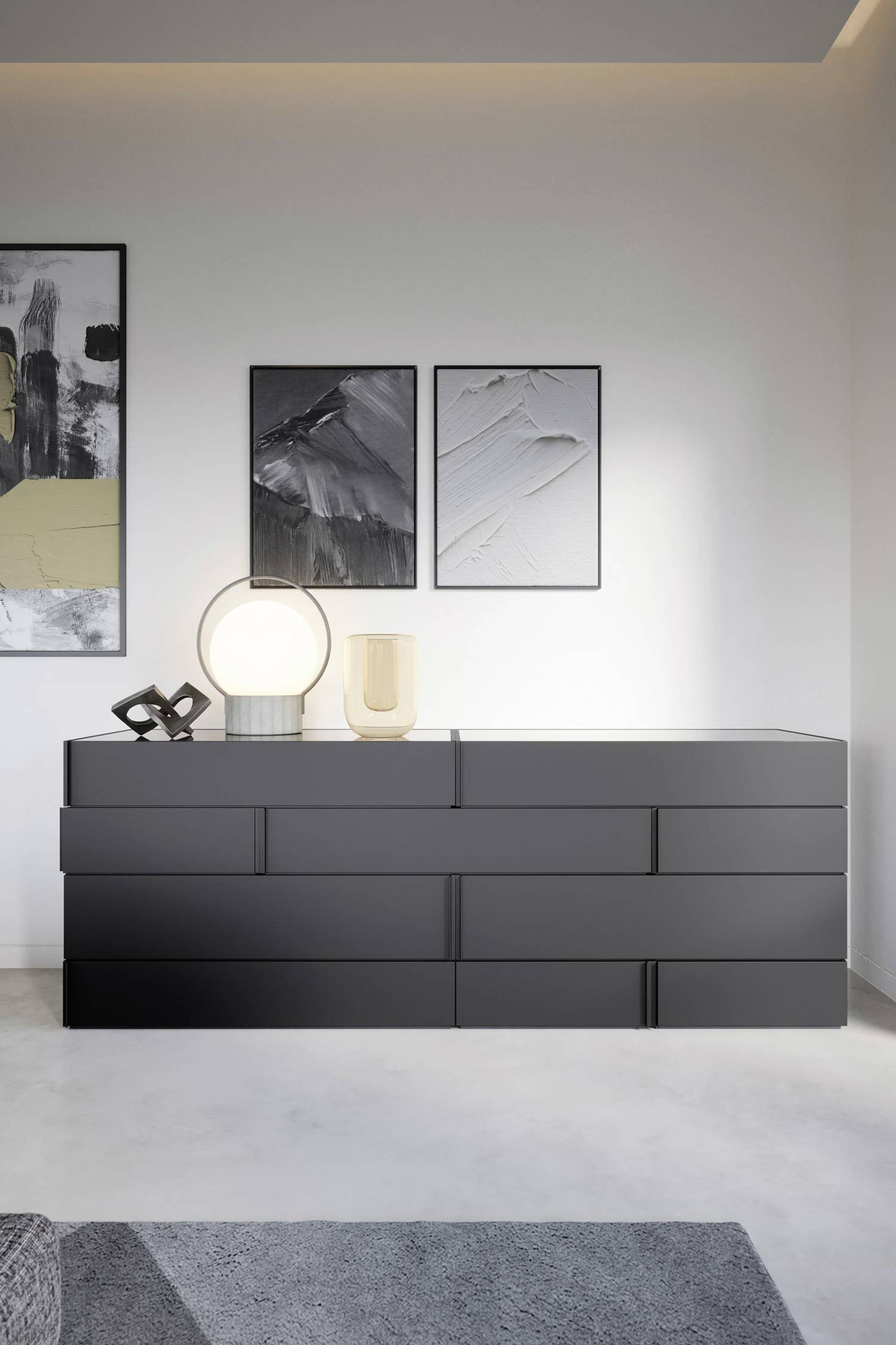 A full-length view of a dark gray drawer unit with an offset, layered drawer face design. The unit is placed against a white wall and is decorated with a round, glowing table lamp and abstract black, white, and gold artwork.
