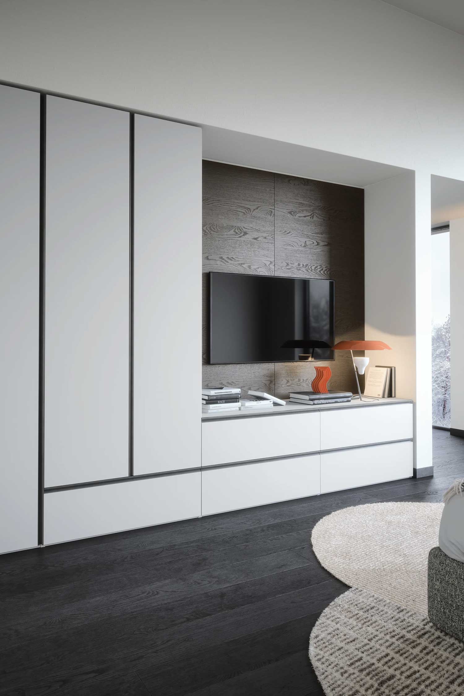 A modern, white wardrobe with vertical black handles. The wardrobe features an integrated media center with a mounted television and a wooden accent wall. The floor is dark wood, and a round rug is in the foreground.