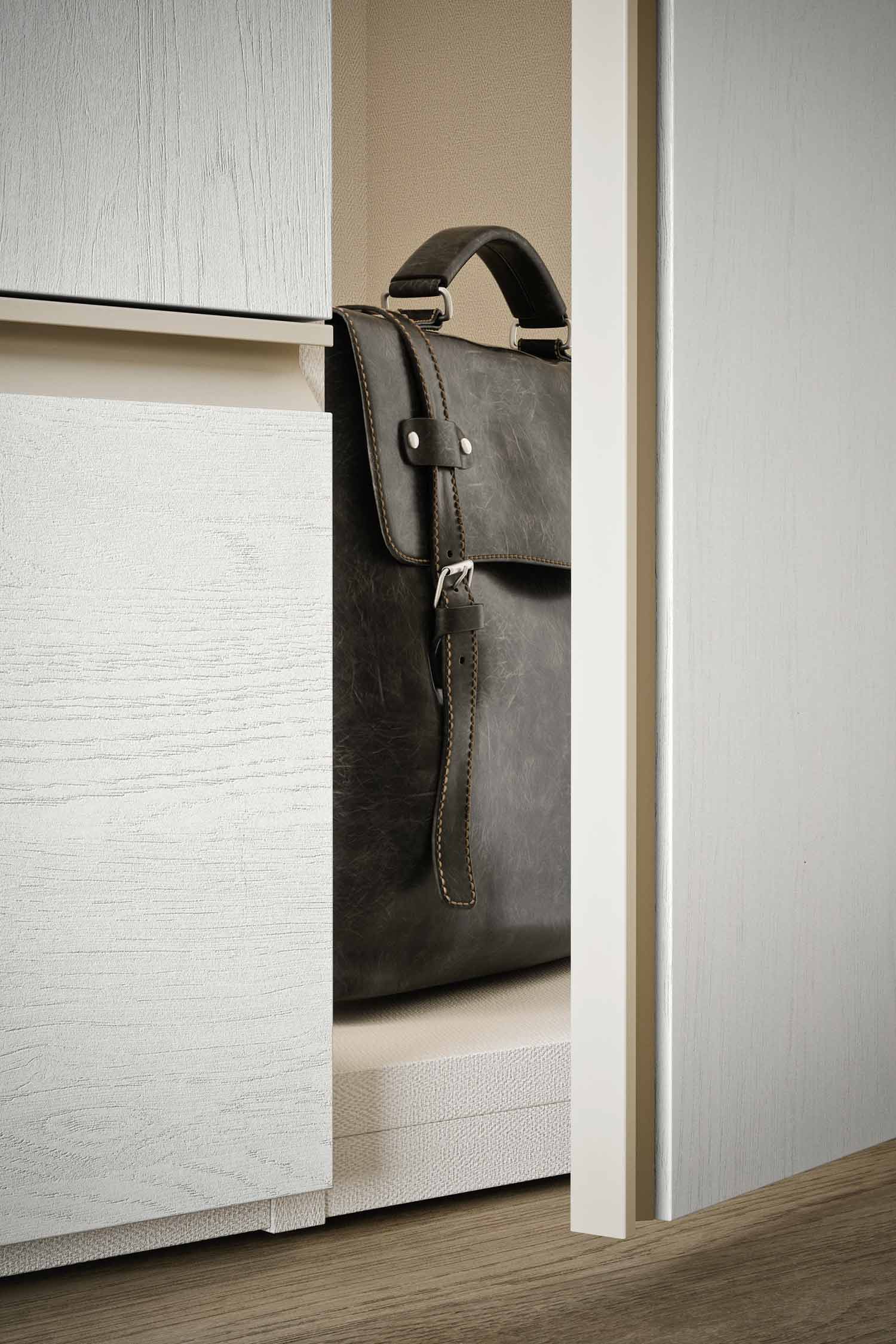 A detailed shot of an open wardrobe with a textured, light wood finish. A large, dark leather briefcase sits on a shelf inside the wardrobe.