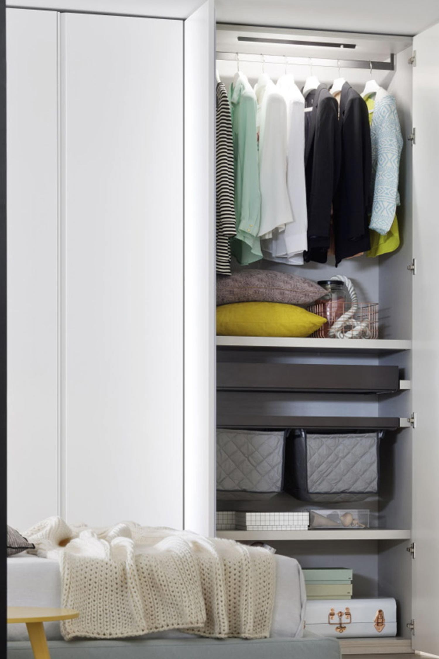 A close-up view of the interior of a custom white wardrobe with a light gray interior. The wardrobe has built-in features including a clothes rack with hanging garments, shelves, and storage baskets.