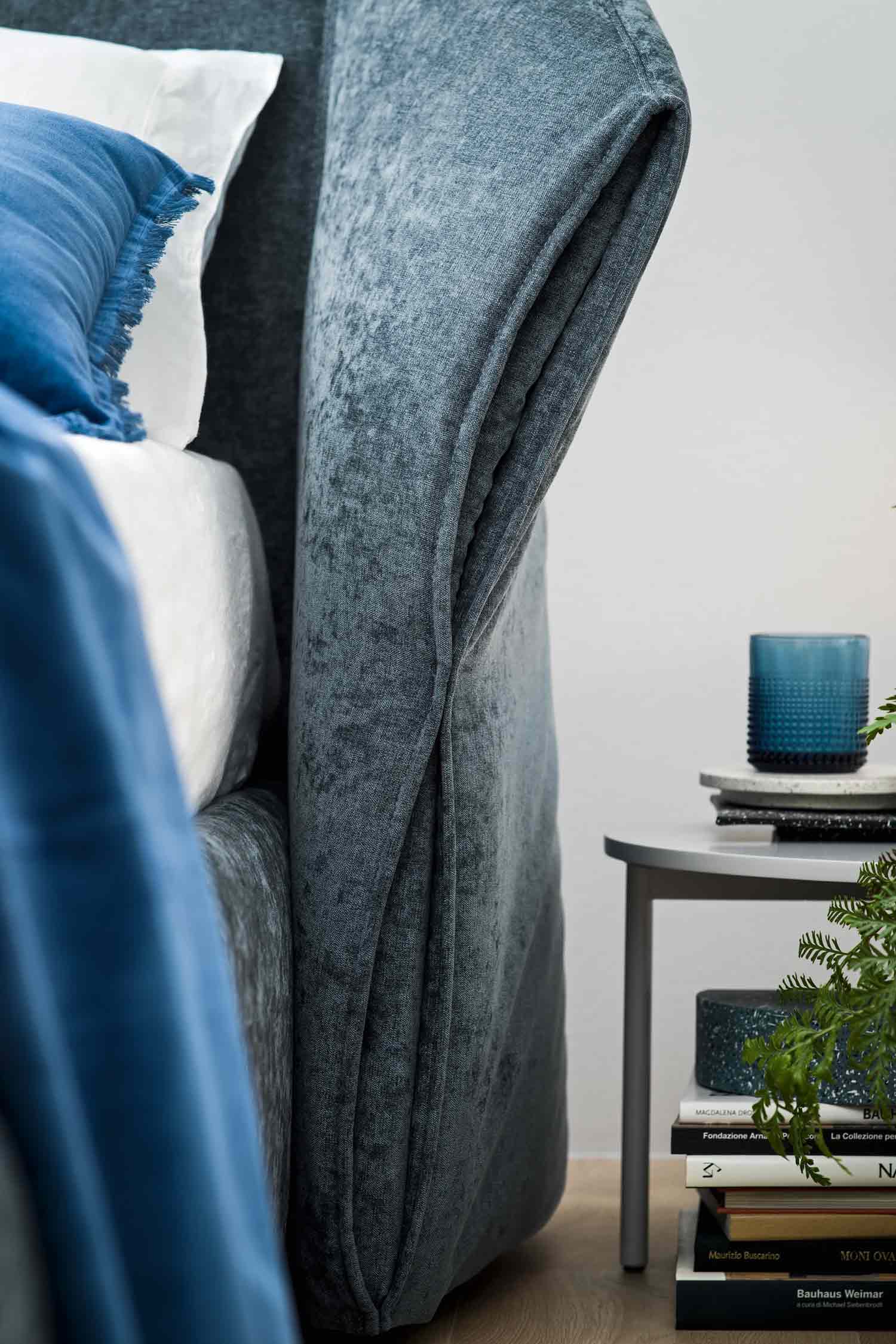 Close-up detail shot of the soft, draped, velvet-like gray-blue upholstery on the headboard of the "Hide" bed, with a side table, books, and a glass candle holder visible next to it.