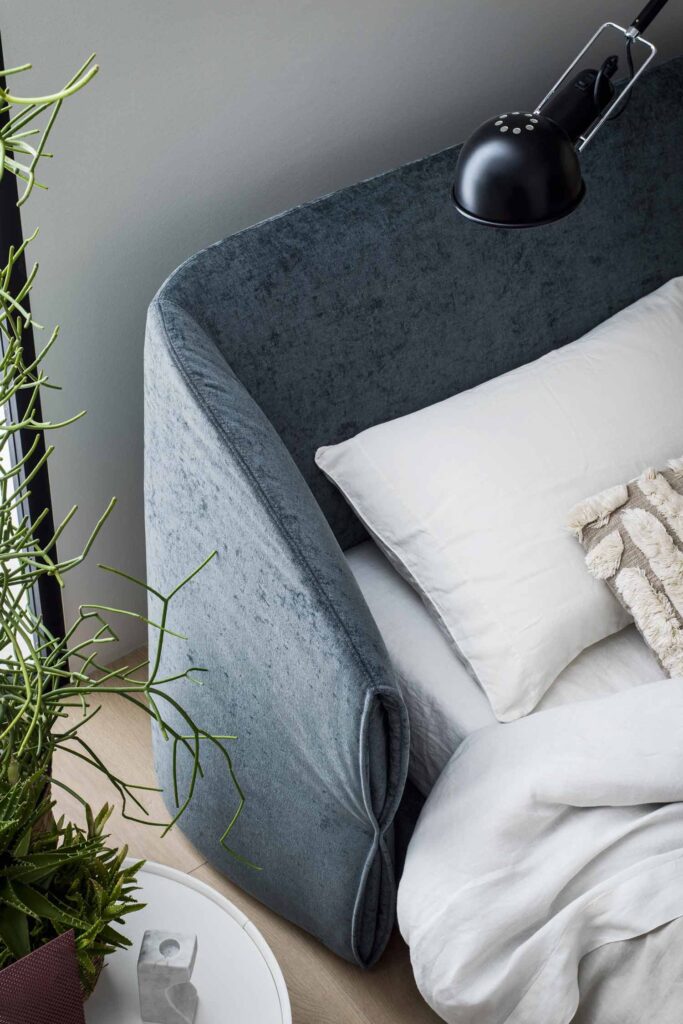 Overhead close-up of the curved, plush dark blue upholstered headboard and side rail of the "Hide" bed, showing white bedding and a black adjustable task lamp mounted above.