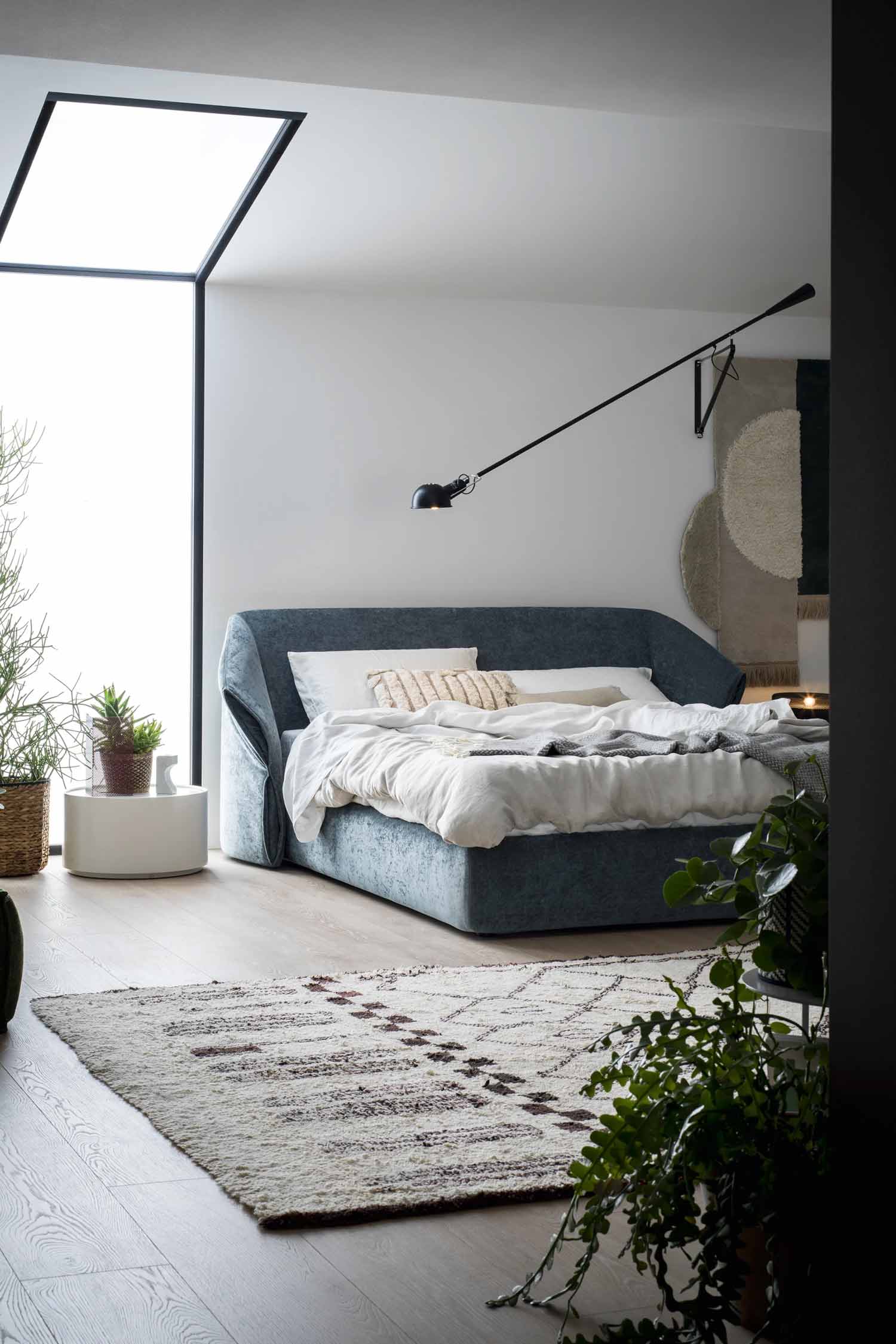 Stylish bedroom featuring the "Hide" luxury bed with a curved, dark blue velvet headboard, light bedding, a textured rug on light wood flooring, a large corner window providing natural light, and several indoor plants.