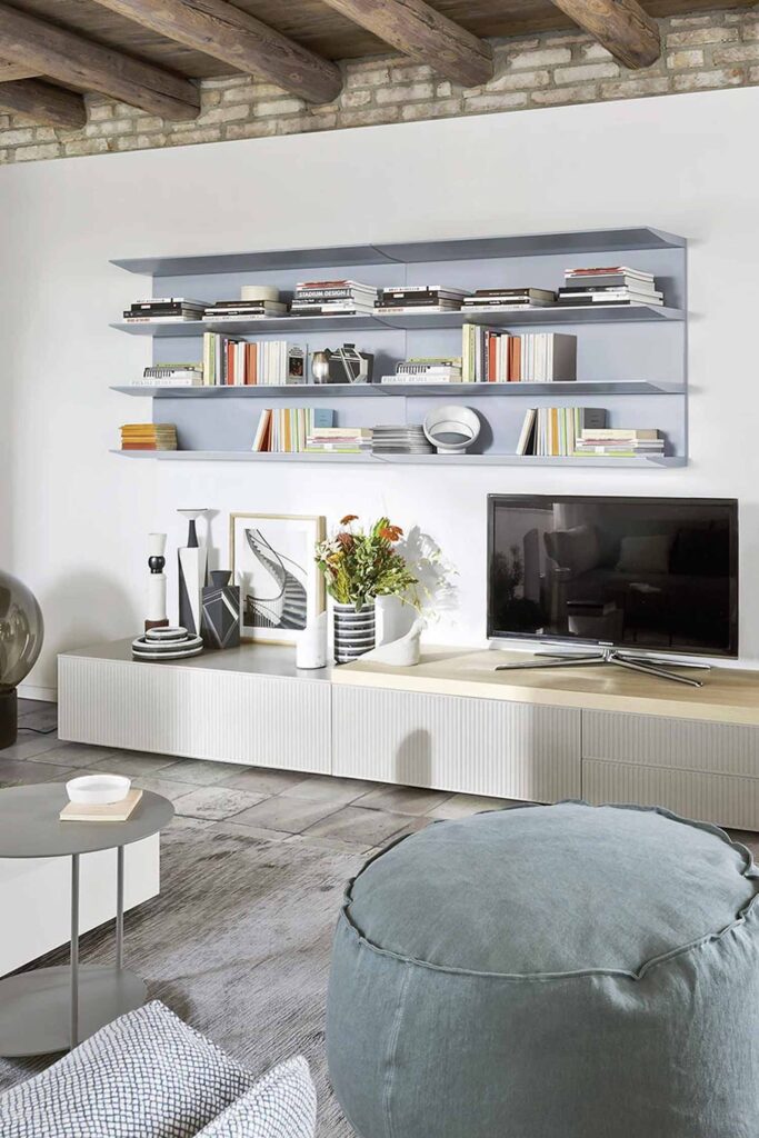 A well-lit modern living room features a long, light-colored media console with a television to the right. Above the console, three light gray floating shelves are filled with neatly organized books and decorative items. Various decorative objects, including vases and framed artwork, are arranged on the media console. In the foreground, a large, round, light blue fabric pouf rests on a textured gray floor, and a small, round gray coffee table is visible to the left. The room has a white wall on the left and a visible brick wall section on the upper right, with exposed wooden beams on the ceiling.