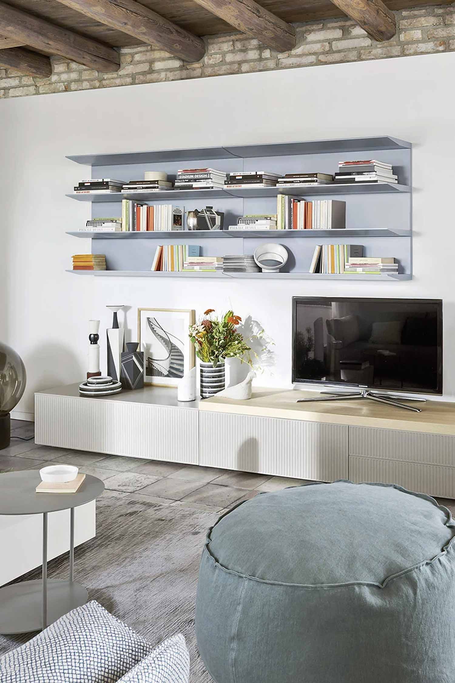 A well-lit modern living room features a long, light-colored media console with a television to the right. Above the console, three light gray floating shelves are filled with neatly organized books and decorative items. Various decorative objects, including vases and framed artwork, are arranged on the media console. In the foreground, a large, round, light blue fabric pouf rests on a textured gray floor, and a small, round gray coffee table is visible to the left. The room has a white wall on the left and a visible brick wall section on the upper right, with exposed wooden beams on the ceiling.