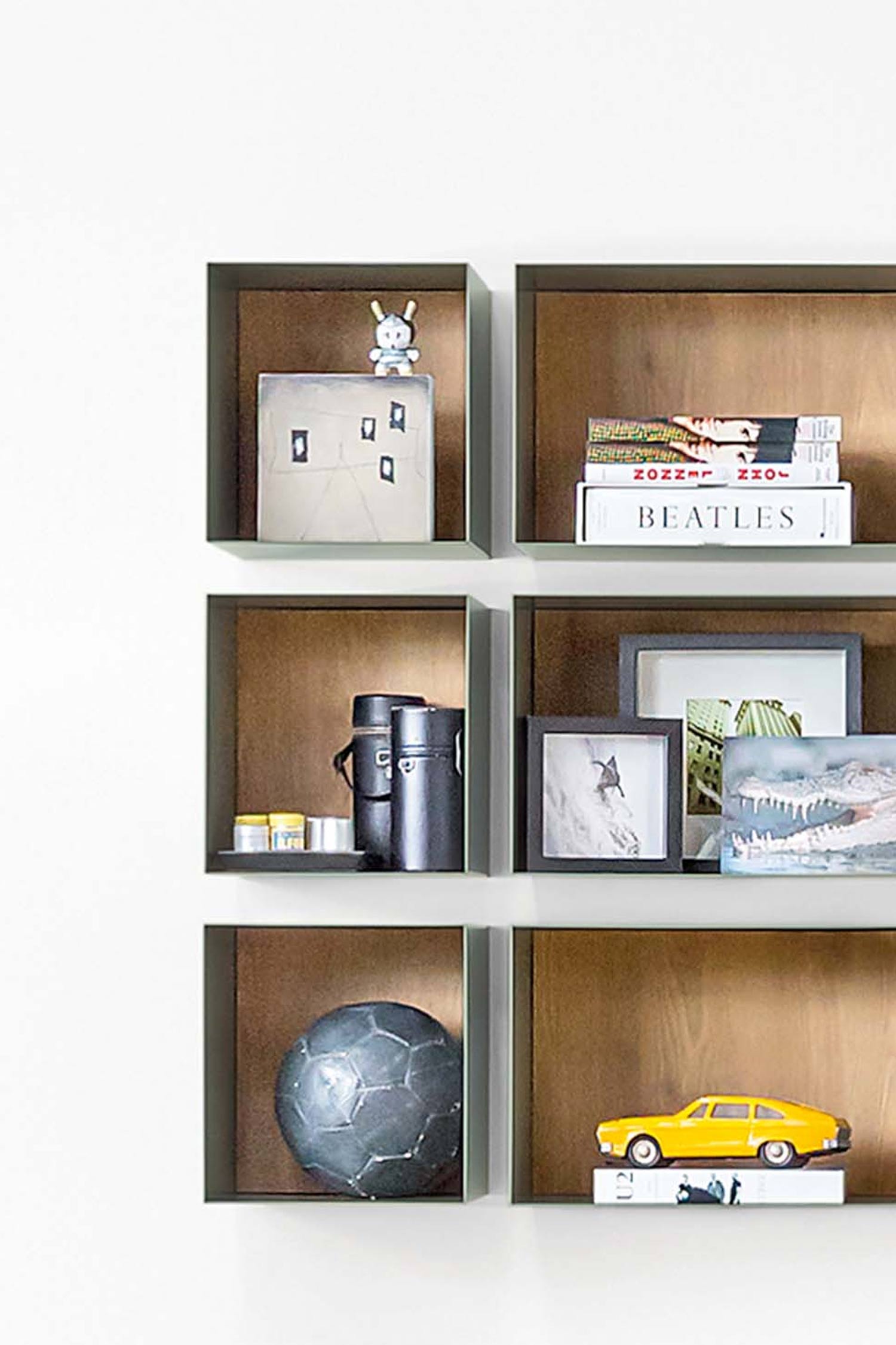 A close-up of several olive green wall-mounted cube shelves with natural wood-toned interiors. The top left shelf holds a light-colored decorative box with a small white bunny figurine. The top right shelf has a stack of books, including one titled "BEATLES." The bottom left shelf holds a dark, spherical, textured object, possibly a decorative ball. The bottom right shelf features a bright yellow toy car resting on books.