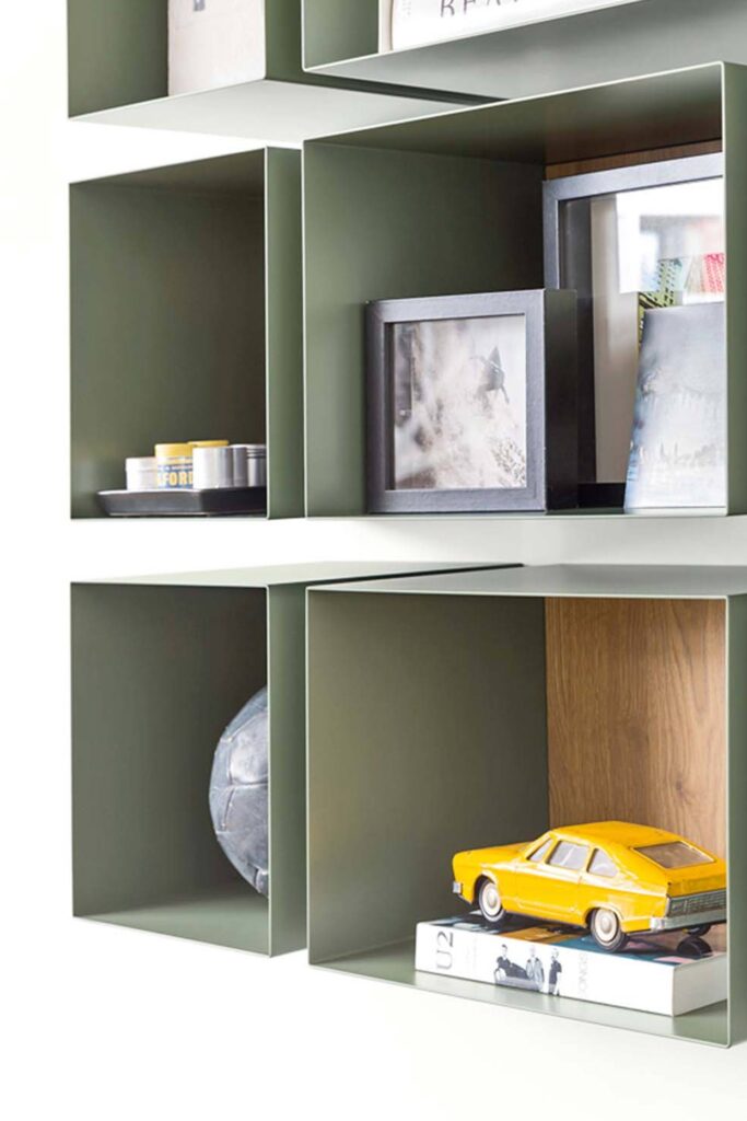 A close-up of several olive green wall-mounted cube shelves with natural wood-toned interiors. The middle left shelf contains two dark cylindrical objects and some smaller containers. The middle right shelf displays framed pictures. The bottom left shelf holds a dark, spherical, textured object, possibly a decorative ball. The bottom right shelf features a bright yellow toy car resting on books.