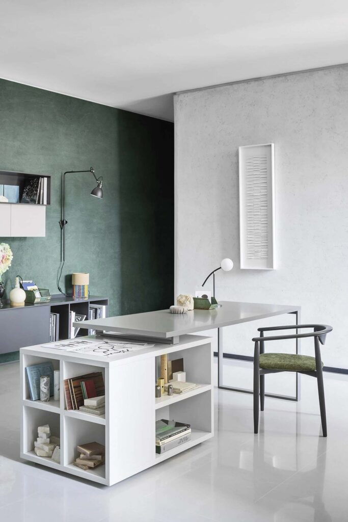 A full-height shot of a modern home office. A long, grey floating desk extends from a white bookshelf unit with books and objects. A black chair with a green fabric seat is positioned in front of the desk. In the background, a dark green accent wall with a wall-mounted light and a grey floating cabinet contrasts with a textured white wall featuring a long, vertical piece of white abstract artwork. The floor is a polished light-grey tile.