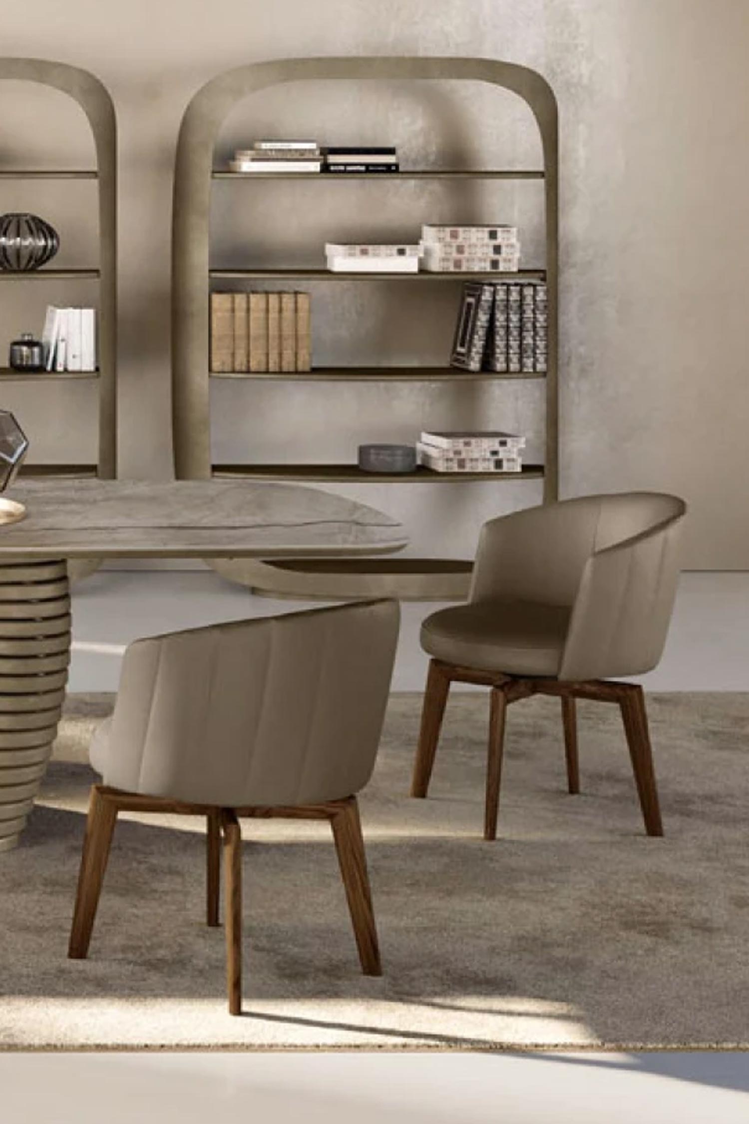 A close-up view of two taupe upholstered dining chairs with wooden legs, positioned around a contemporary dining table with a sculptural base. One chair is facing away, showing the vertically stitched backrest, while the other faces forward. In the background, two tall, arched bookshelves are filled with decorative items and books.