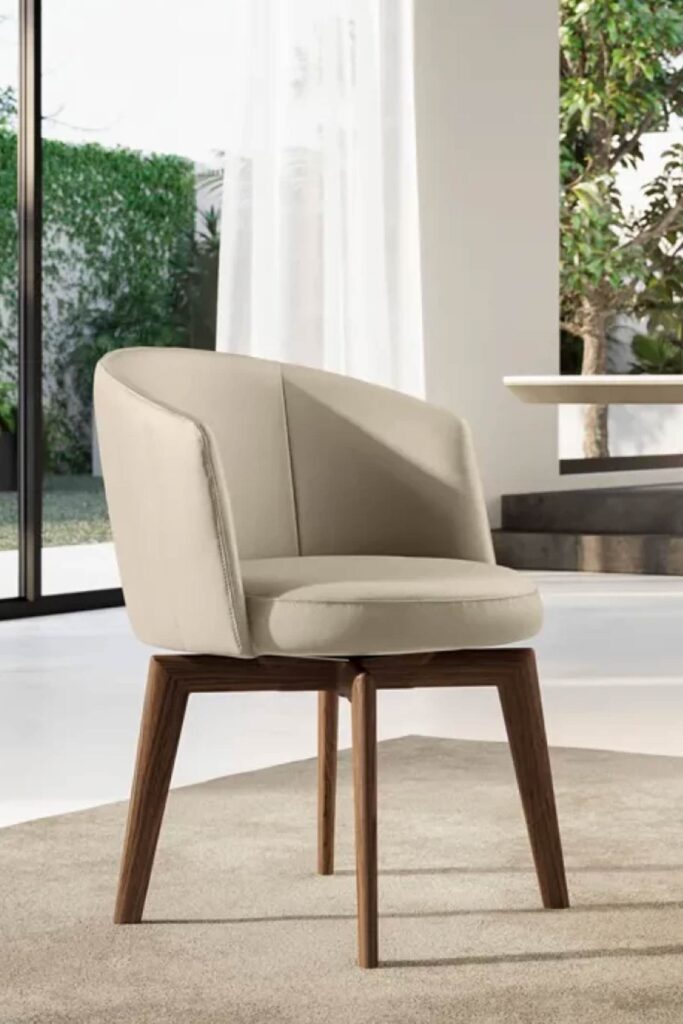 Detailed view of a single luxury dining chair with a light beige leather seat and back, featuring decorative stitching. The chair sits on dark wooden legs on a light rug, next to a large sliding glass door that opens to a sunny, enclosed outdoor garden.