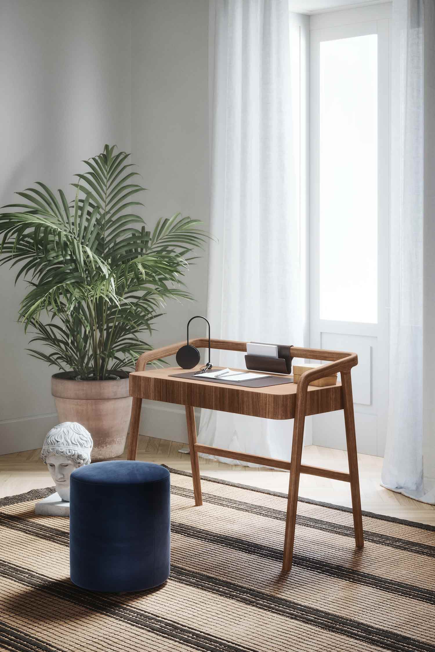 A contemporary wooden desk with a brown leather desk pad is positioned in a sunlit room. On the desk, there is an open notebook, a black lamp, and a brown leather folder. To the left of the desk, a large potted plant and a white classical bust are visible. A blue velvet stool sits in front of the desk, all placed on a striped woven rug. A large window with sheer white curtains is on the right side of the image.