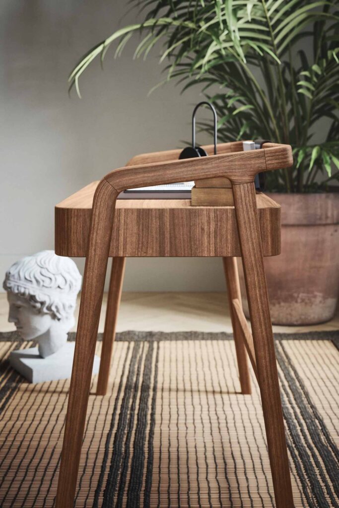 A close-up, low-angle shot of a modern wooden desk with a minimalist design. The desk features slender legs and a curved backrest. On the desk surface, a black desk lamp and some papers are visible. To the left, a white classical bust rests on the floor, and in the background, a large potted plant is visible. The floor is covered with a striped woven rug.
