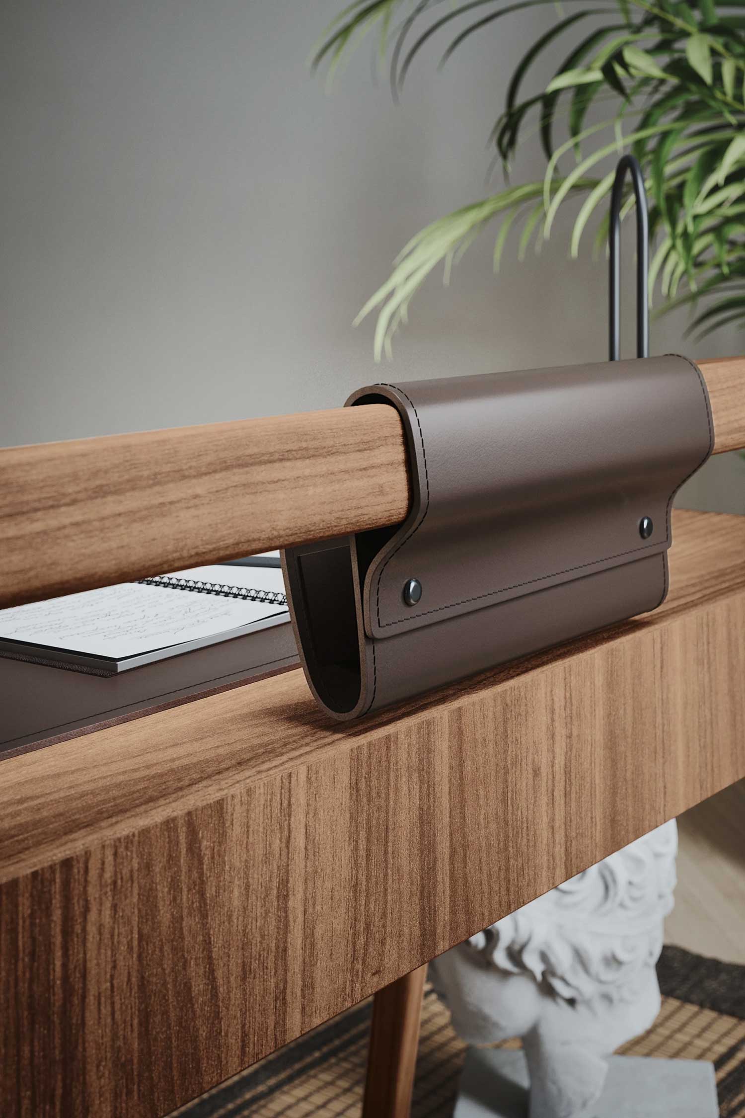 A close-up of a modern wooden desk. A brown leather folder is draped over the top wooden rail of the desk's frame, held in place by two black metal buttons. The wooden desk surface below shows a fine grain, and a notebook and a white classical bust are visible in the soft-focus background.