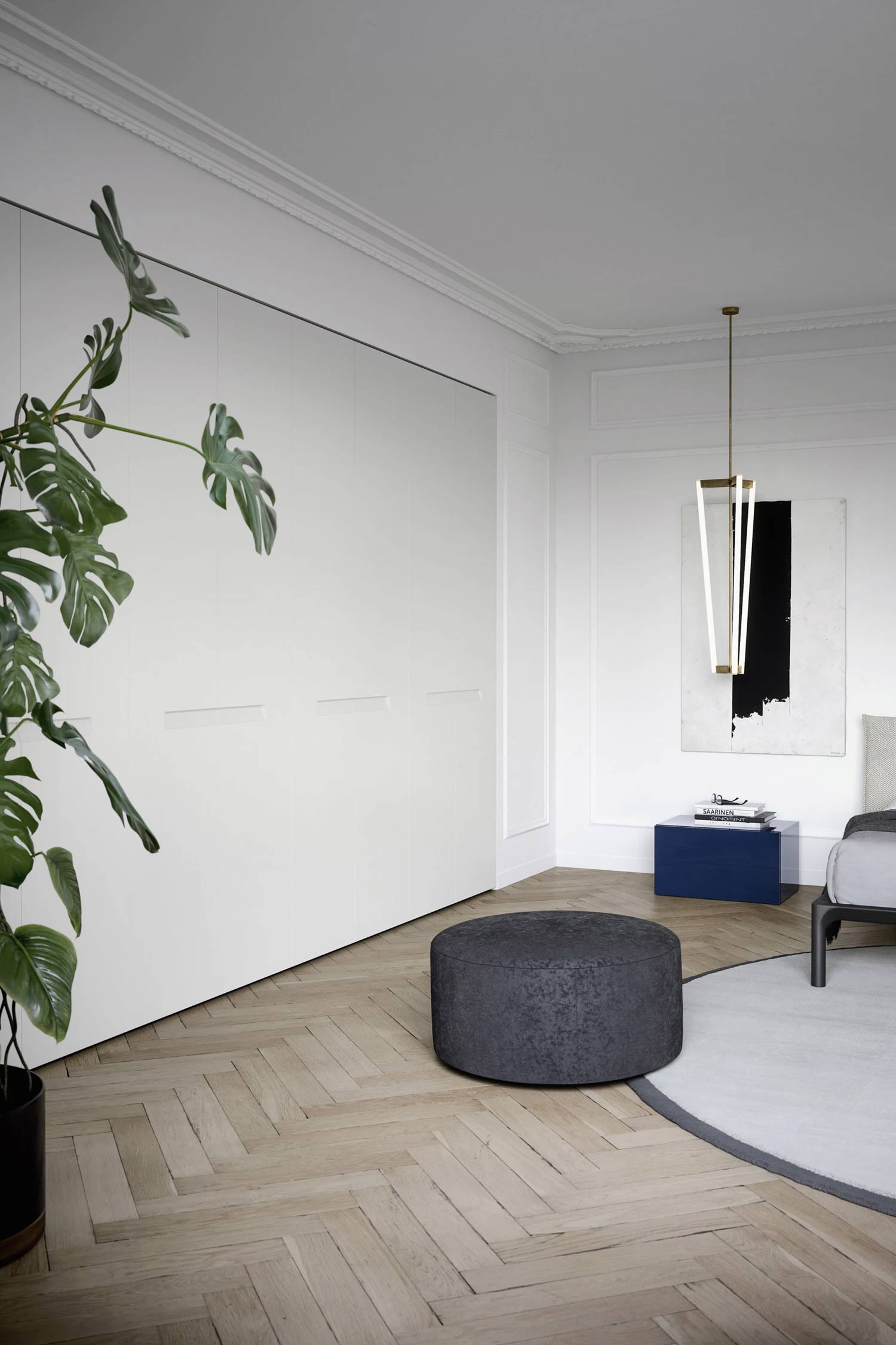 A full view of a modern bedroom with a large, white, floor-to-ceiling wardrobe with a black base. The wardrobe has recessed handles and fills the left side of the room. A gray round pouf, a large potted plant, and a bed are also visible. The room has a hardwood herringbone floor and a black-and-white art piece on the wall.