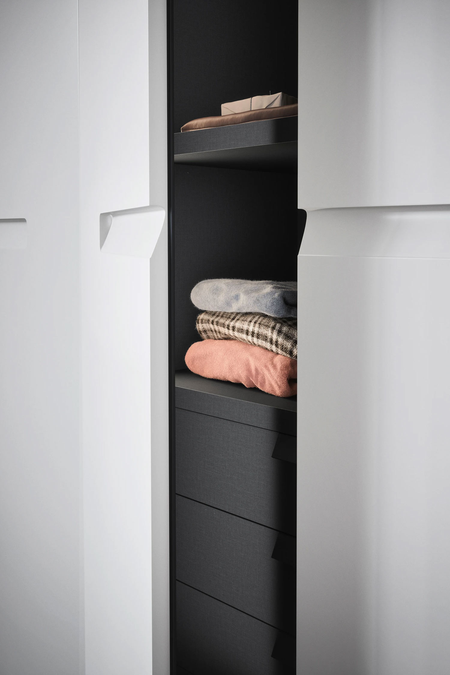 A close-up shot of a white wardrobe with two doors partially open. Inside, a dark gray interior is revealed, with two pull-out drawers and a shelf holding a stack of folded sweaters and a small box. The handles on the inside of the doors are also visible.