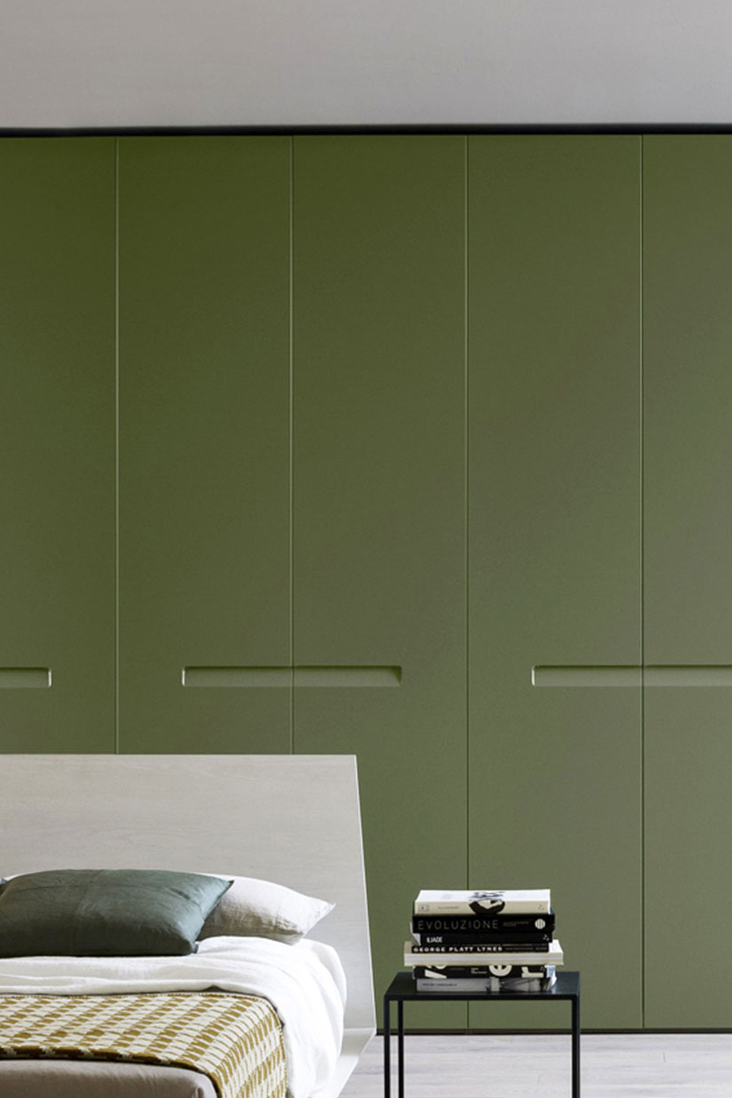 A close-up of a modern, built-in wardrobe with a matte olive green finish. The six doors have recessed handles, and the bottom is flush with the floor. A bed with a light gray headboard and a small black side table with books are in the foreground.