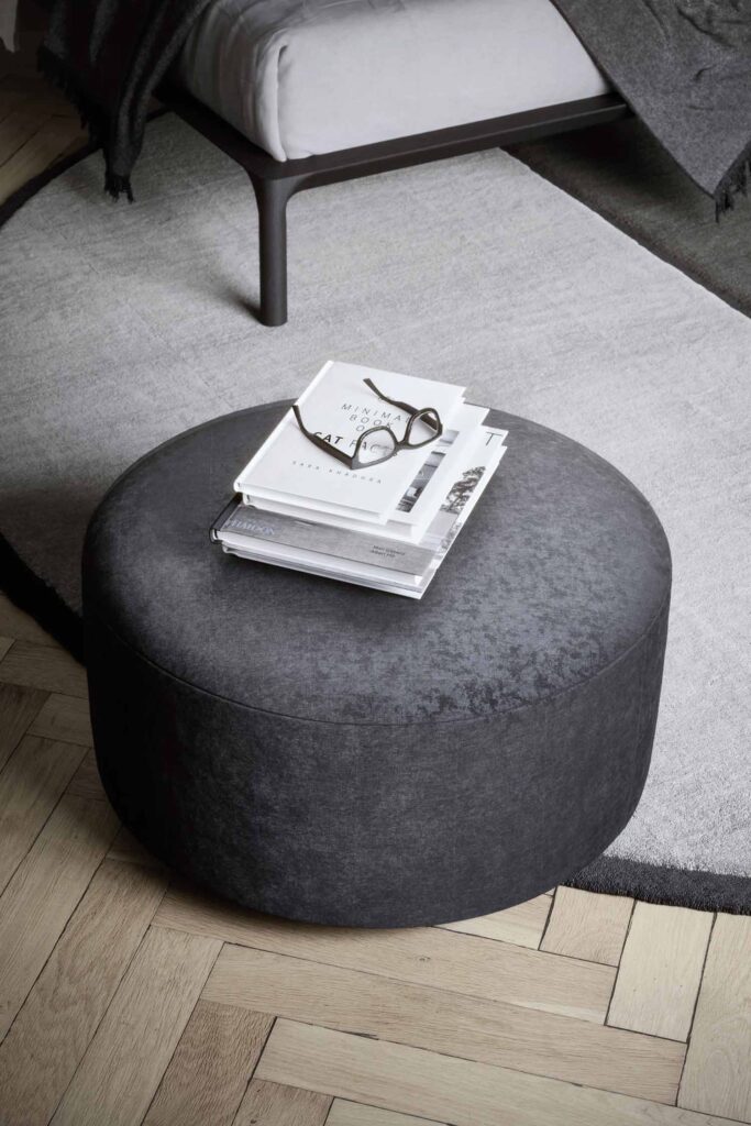 A close-up, high-angle shot of a round, dark gray ottoman with a slightly mottled texture. On top of the ottoman, there is a stack of books with a pair of eyeglasses resting on the top book. The ottoman sits on a light gray rug with a dark border, and the flooring is a light wood herringbone pattern. In the upper-left corner, the corner of a bed frame and a dark gray blanket are visible.