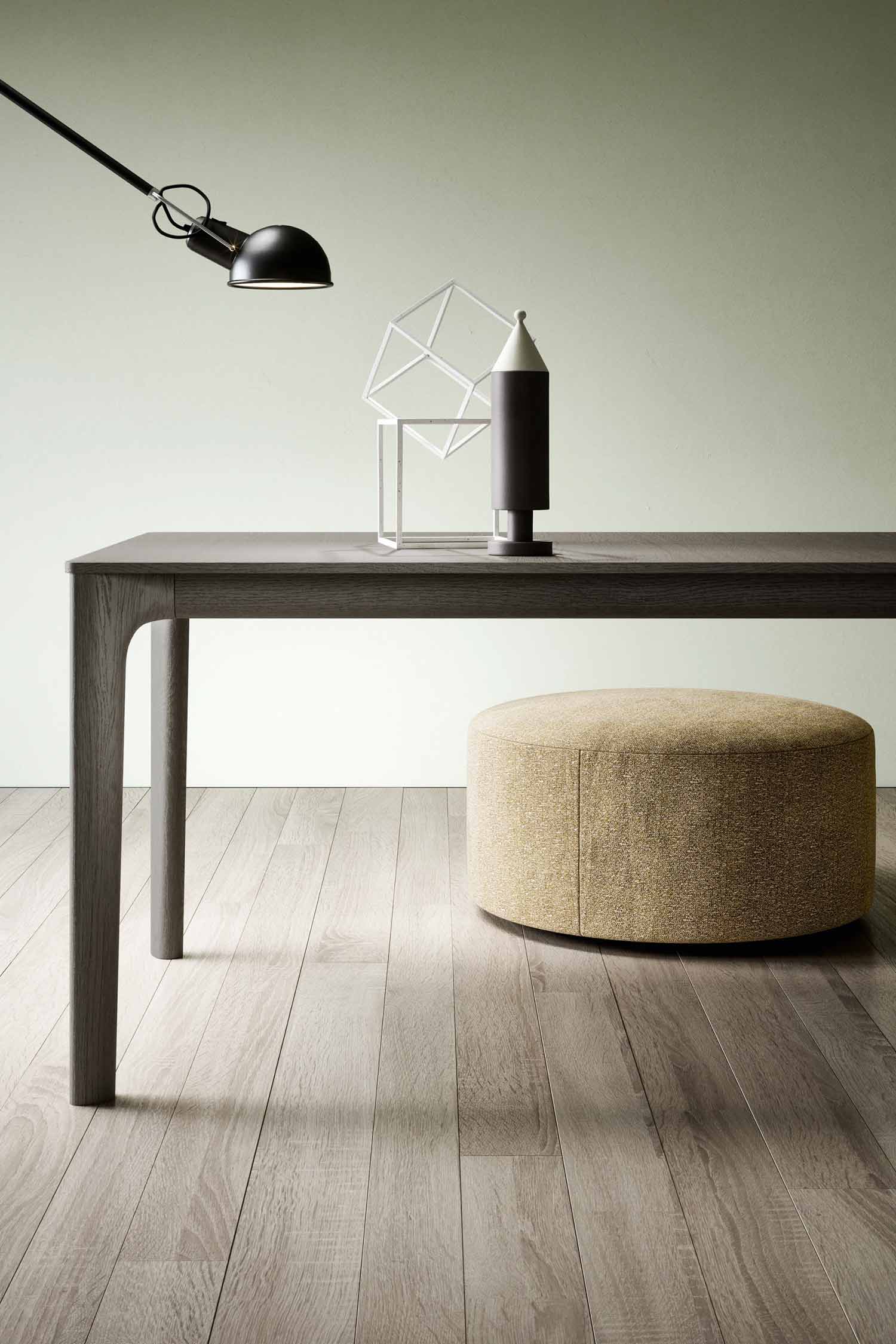 A close-up view of a dark wood table with a light, textured yellow-beige ottoman tucked underneath. A black and white decorative object with a geometric wire frame sits on the table's surface. A black industrial-style lamp is mounted on the wall to the left, extending over the table. The flooring is a light-colored wood, and the wall in the background is a soft, pale green.