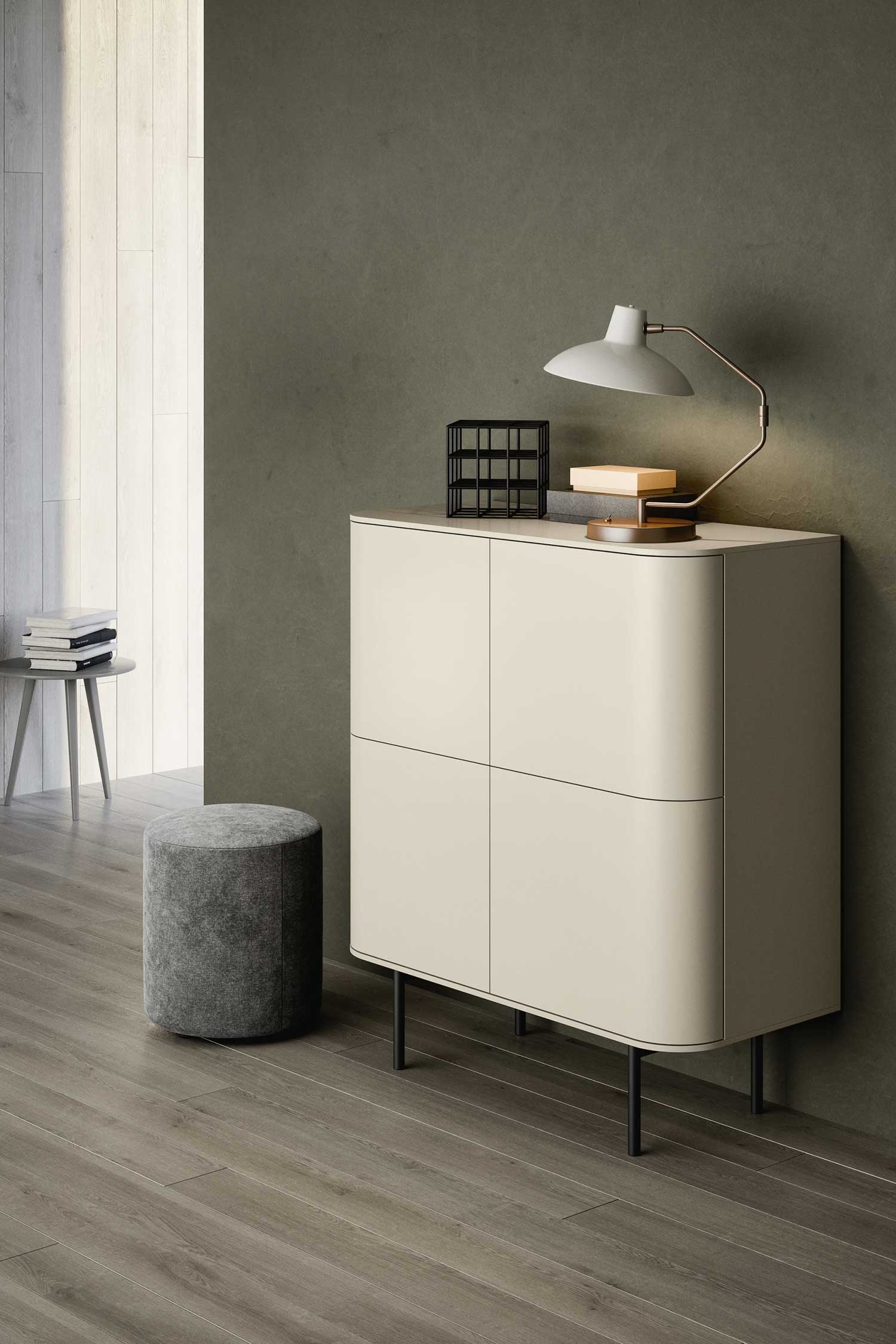 A minimalist room featuring a beige, rounded-corner cabinet with four doors and black legs. A modern white and copper table lamp sits on top, next to a small black decorative grid and a book. To the left of the cabinet, there is a round, gray upholstered ottoman and a small gray side table with a stack of books. The wall behind is a dark gray with a textured finish, and the floor is a wide-plank wood.