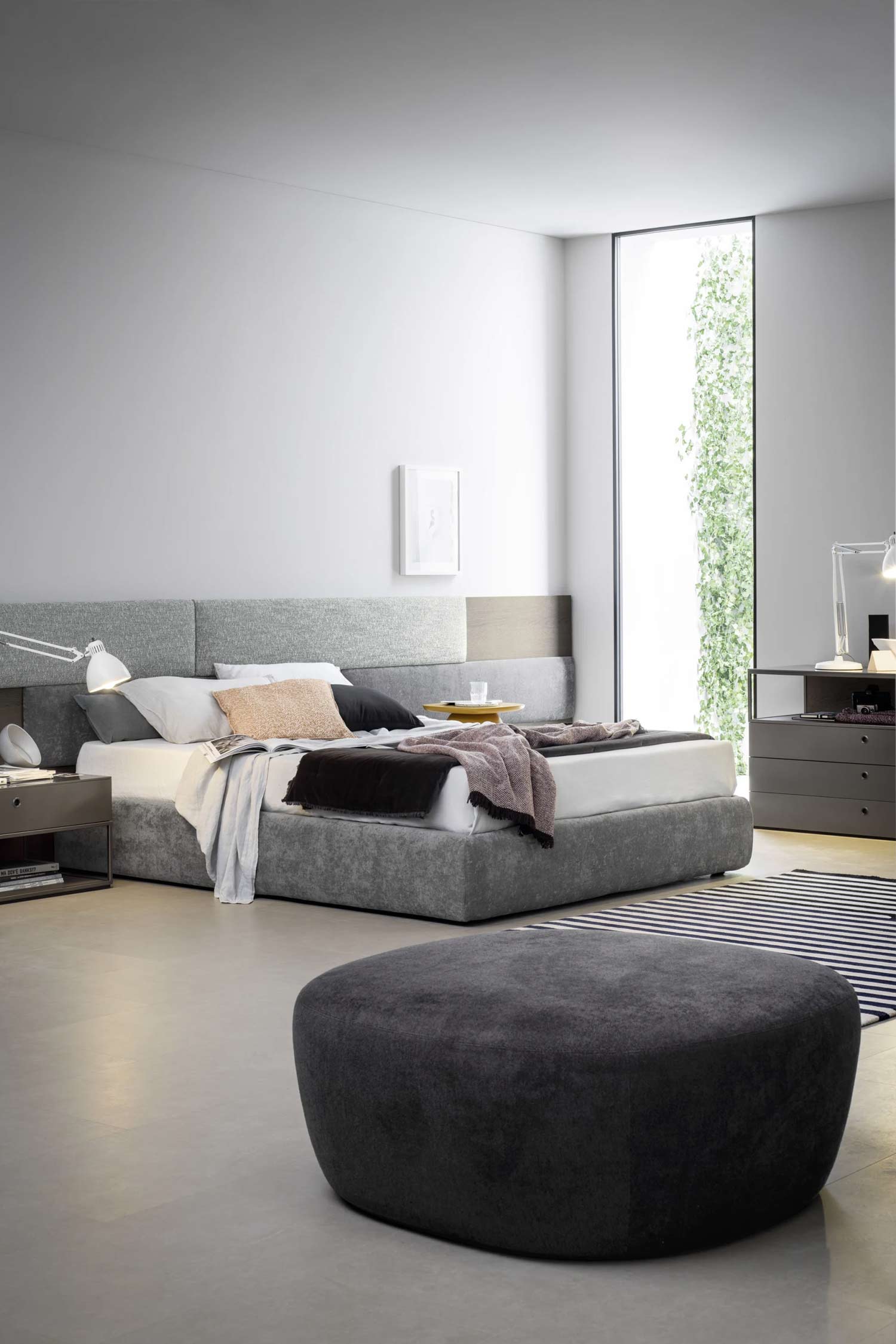 A large, low-profile gray upholstered bed is positioned in a minimalist bedroom. The bed has a unique, gray-paneled headboard and is covered with a variety of pillows and blankets. A small, white task lamp is on the nightstand to the left, and a small yellow tray sits on the bed. A large, dark gray ottoman is in the foreground, resting on a black and white striped rug. The room has light gray walls and a light gray floor, and a tall, narrow window on the right side provides a view of a lush green vine climbing a wall outside. A matching gray dresser with a similar white lamp is visible on the far right.