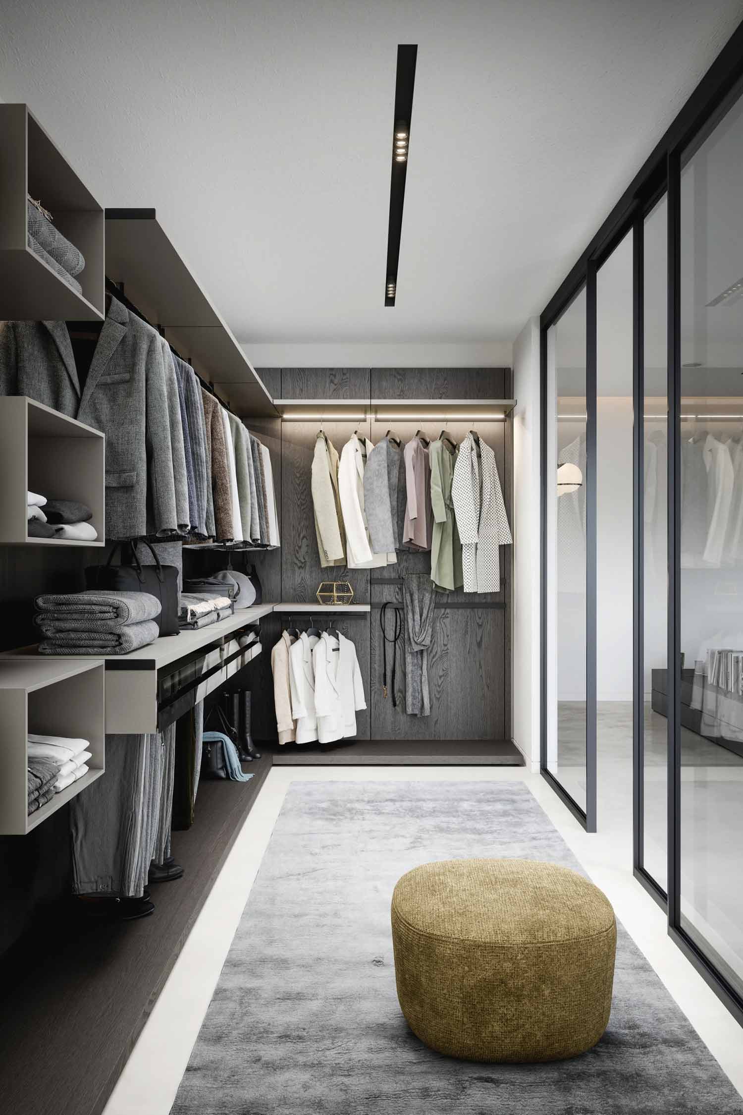 A spacious and modern walk-in closet is shown from a low angle. The closet features dark wood paneling and open shelving on the left, where neatly folded clothes and bags are stored. Hanging rods are filled with a variety of blazers, coats, and blouses. On the floor, a textured, light brown, rounded ottoman sits on a plush gray rug. The room is well-lit, with track lighting on the ceiling and a glass partition on the right, which leads into another part of the bedroom.