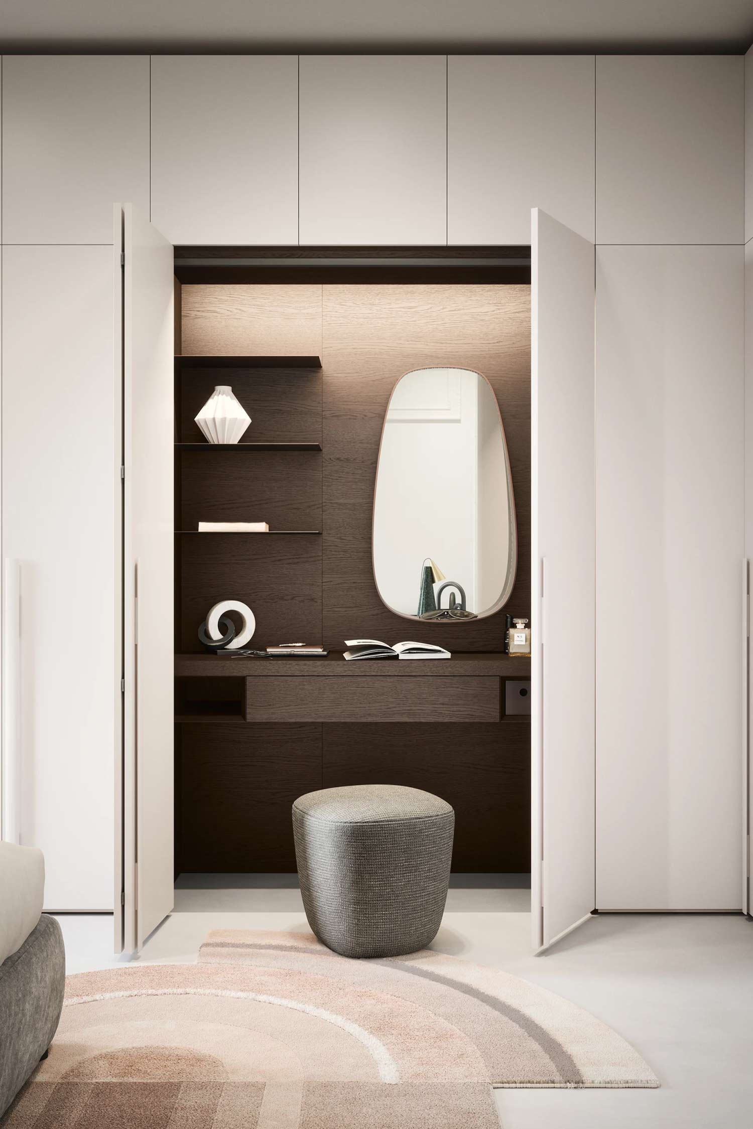 A compact and modern built-in vanity is nestled between two white wardrobe doors. The vanity features dark wood with warm, recessed lighting illuminating a light wood back panel. A unique, asymmetrical mirror hangs above the counter, which holds an open book, a lamp, and a few small decorative objects. Open shelves on the left display a white vase and another decorative object. A rounded, upholstered gray ottoman sits on a pink and brown patterned rug in front of the vanity.
