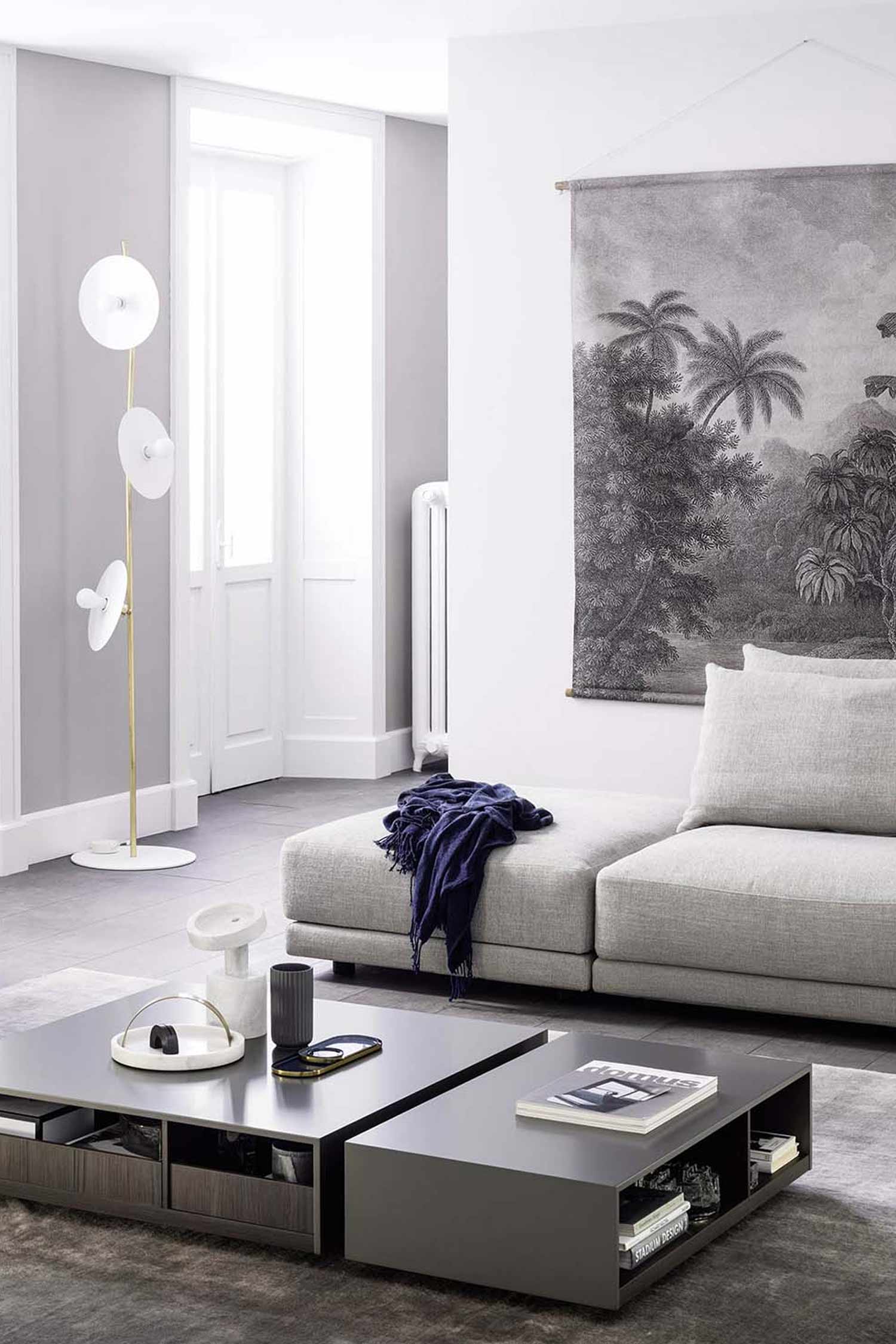 A stylish living room featuring an orange upholstered armchair with a dark wood frame and armrests in the foreground to the left. A textured, light gray ottoman is on a cream-colored rug with a geometric pattern to the right. In the background, a dark wood credenza with vertical grooves is against a white wall. Decorative objects, books, and a unique black wall-mounted lamp are on top of the credenza. The room has light wood parquet flooring, and a large potted plant is visible in the far left corner.