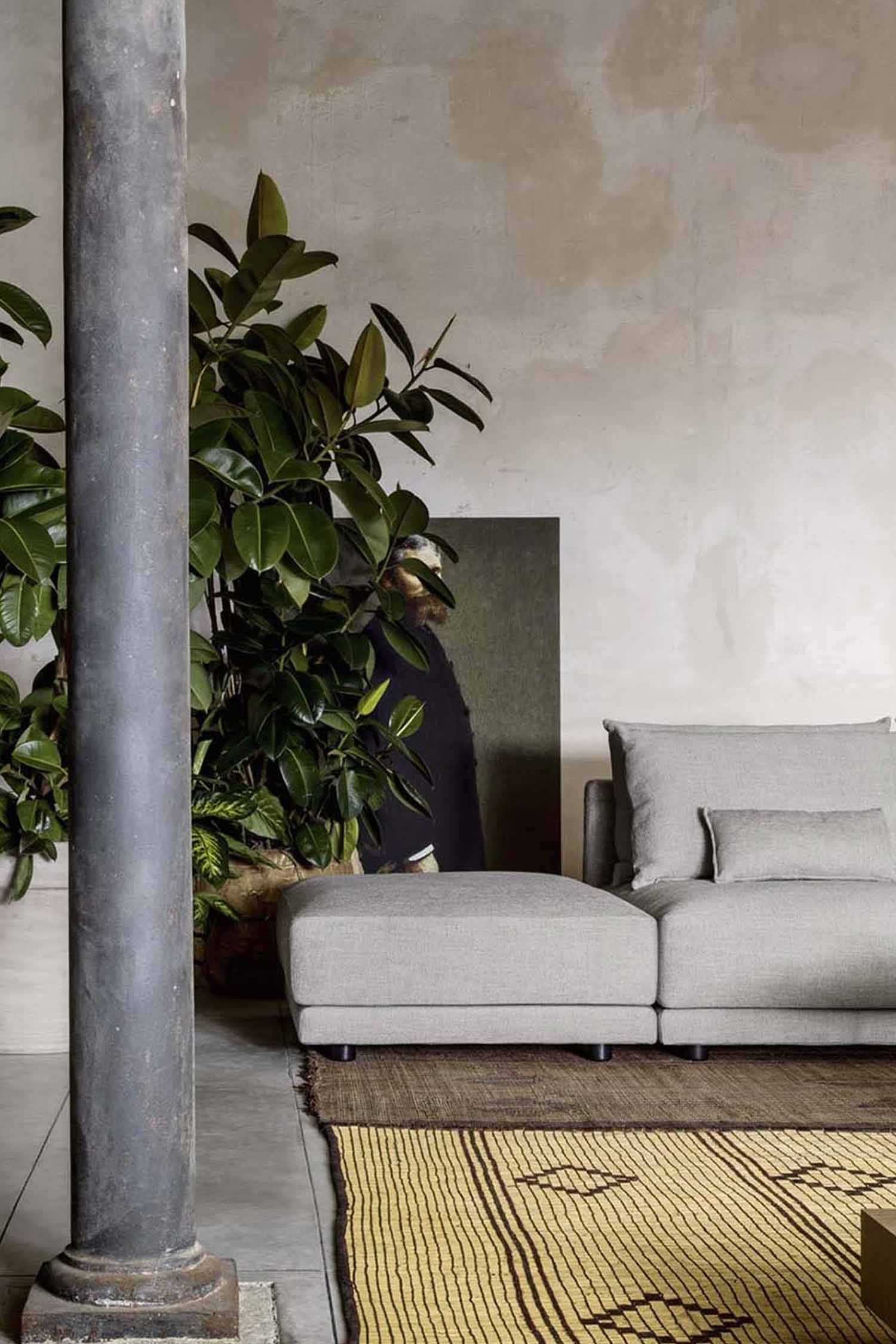 A close-up shot of a modern living room featuring a light gray upholstered ottoman and a matching sectional sofa. A large potted plant with broad green leaves is to the left of the ottoman. In the background, a painting of a man with a beard is partially visible behind the plant. A dark industrial column stands on the far left. The furniture rests on a textured brown rug with a yellow geometric pattern. The wall behind is a rustic, textured white.