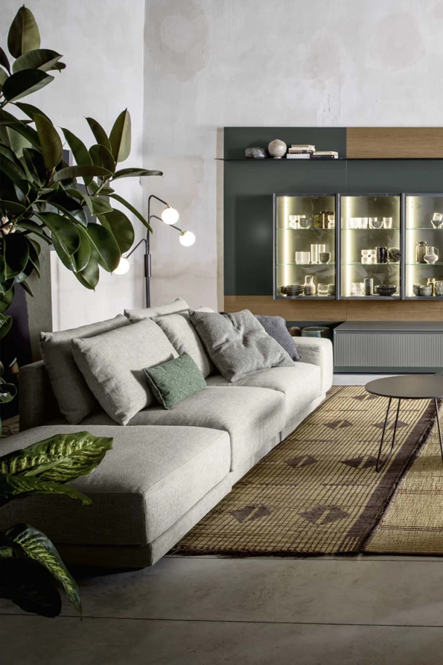A low-angle, wide shot of a spacious and modern living room. A large, light gray sectional sofa with a white cushion and a green pillow is on the left, facing a large, multi-functional wall unit with dark green and light wood panels. The wall unit has a central television, display cabinets with lighting, and various open shelves with books and decorative objects. In the center, a dark gray coffee table with thin metal legs holds a small, red-colored ceramic stool. A bright yellow cylindrical ottoman is in the foreground, next to a large metal support beam. The room has concrete flooring and a textured, tan-colored rug with a geometric pattern. A large potted plant is on the far left and another one is on the far right.