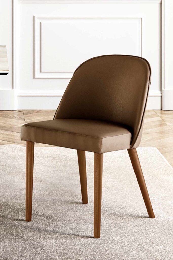 A close-up of a Manta luxury dining chair upholstered in smooth, medium brown leather with light-colored wood legs. The chair is on a light, textured rug in a room with white paneled wainscoting and a wood floor.