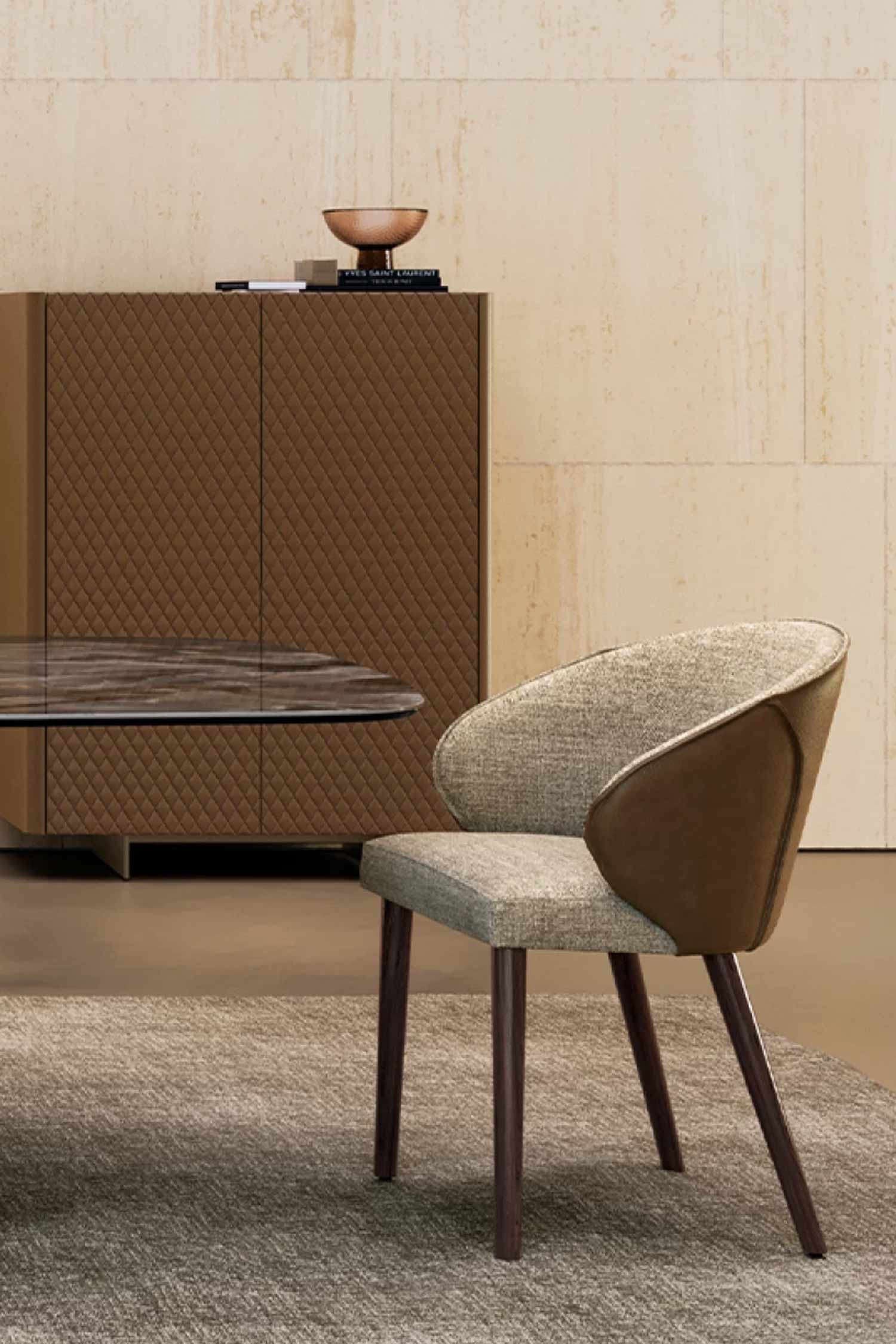 A close-up of a Manta luxury dining chair featuring a light beige tweed fabric seat and a contrasting brown leather accent on the curved back and sides. It is set against a textured, quilted brown cabinet and neutral wall in a dining room.
