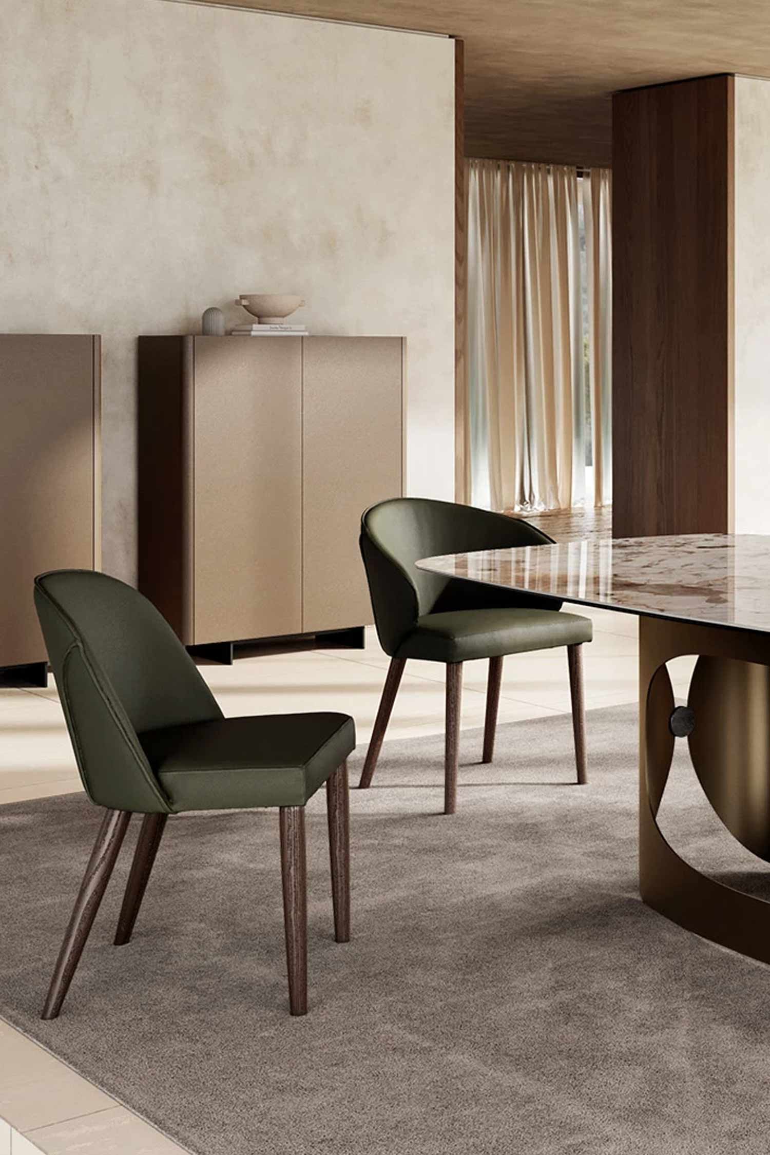 Two Manta luxury dining chairs upholstered in dark olive green leather with dark wood legs are positioned near a glass and marble-top dining table with a bronze base. The chairs are in a modern dining room with neutral-toned cabinetry and sheer curtains.